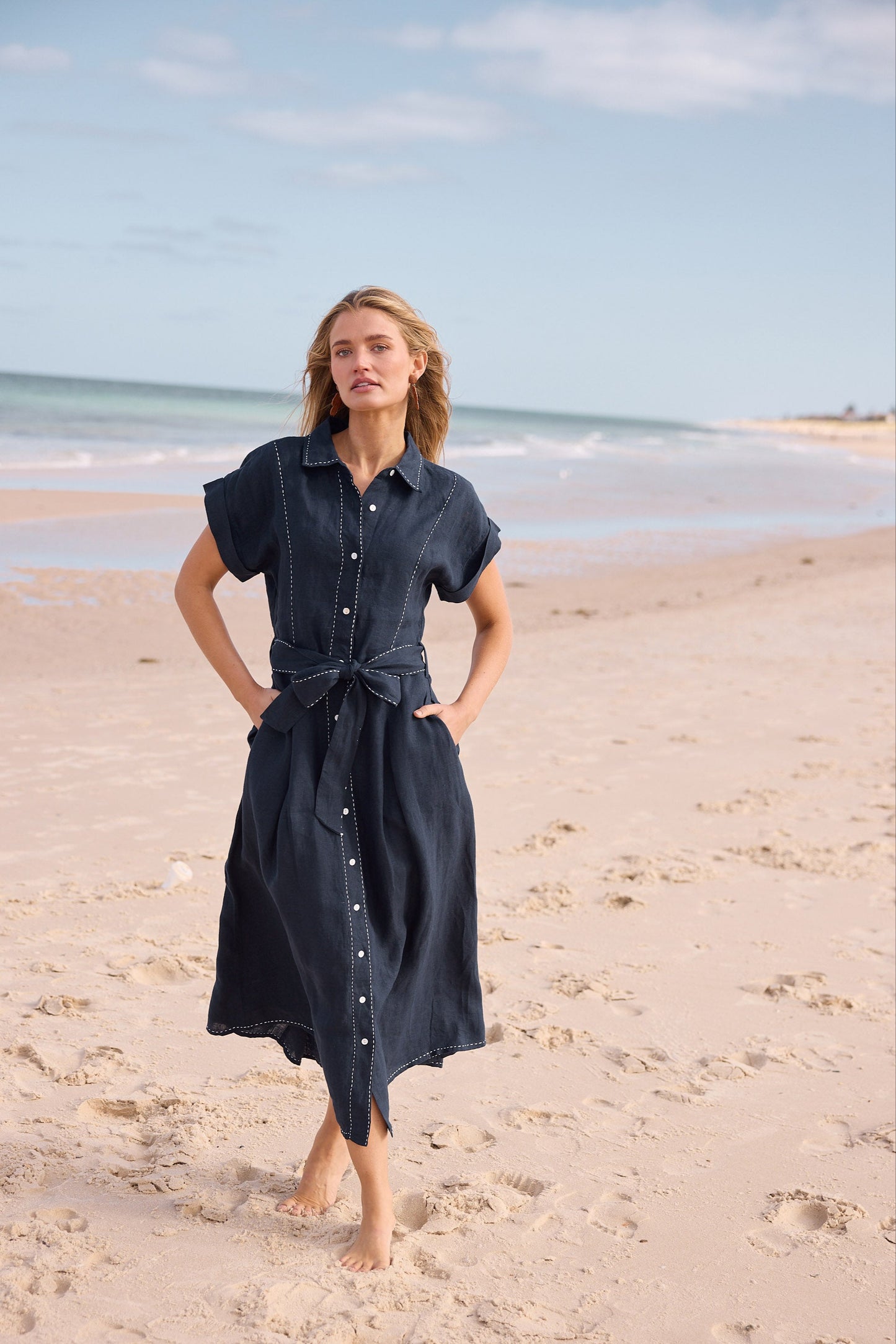 Woman in a dark blue dress standing on a sandy beach with ocean and sky in the background