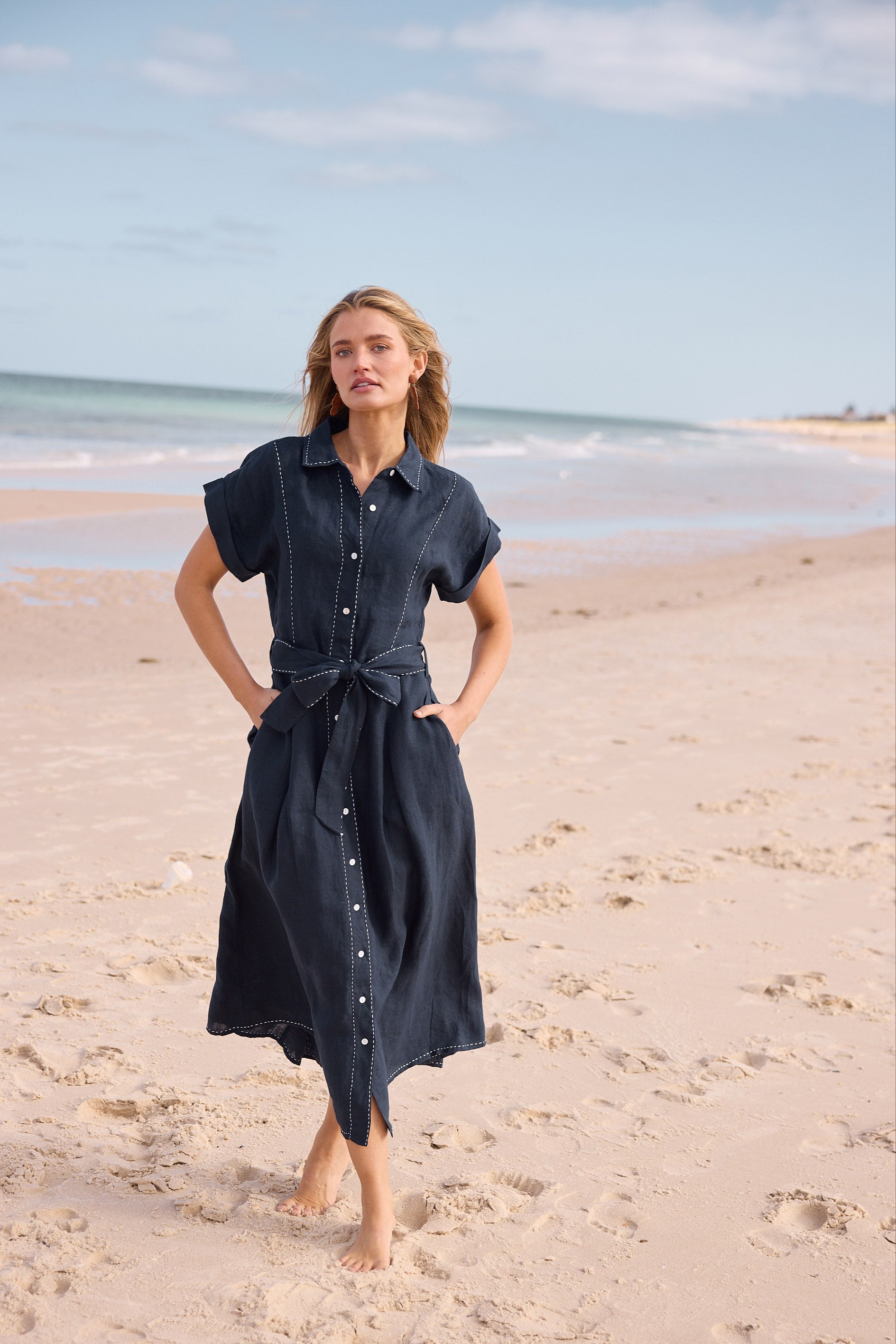 Woman in a dark blue dress standing on a sandy beach with ocean and sky in the background