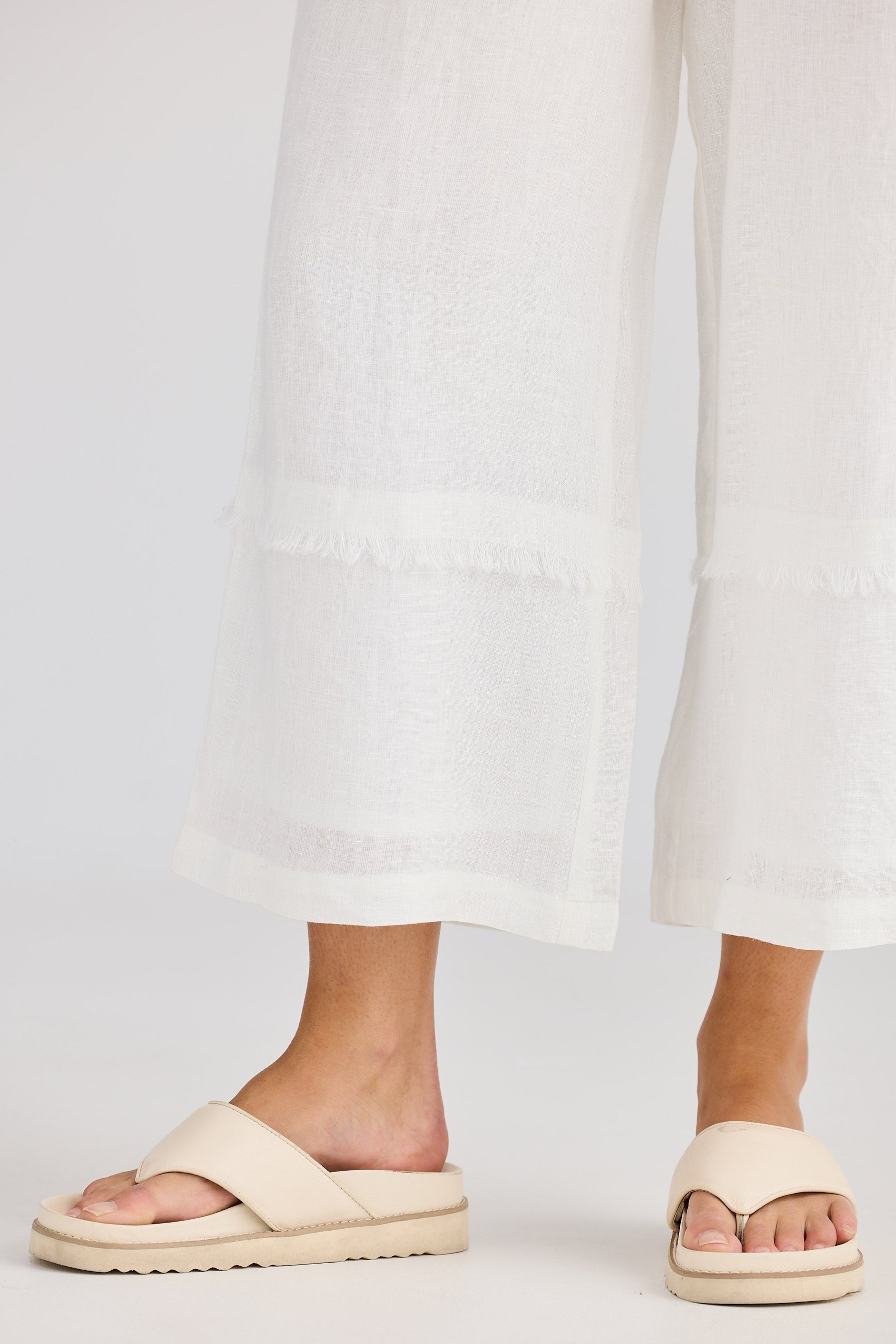 Beige sandals worn with white pants on a light gray background