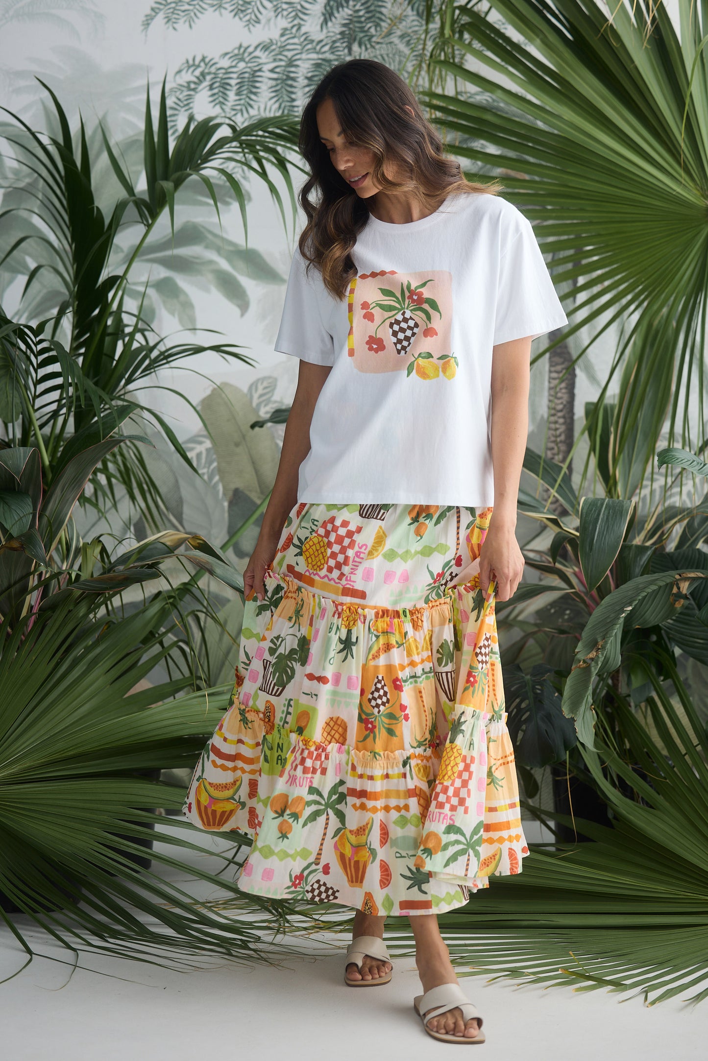 Woman wearing a white t-shirt with a colorful fruit design and a matching skirt, standing among tropical plants.