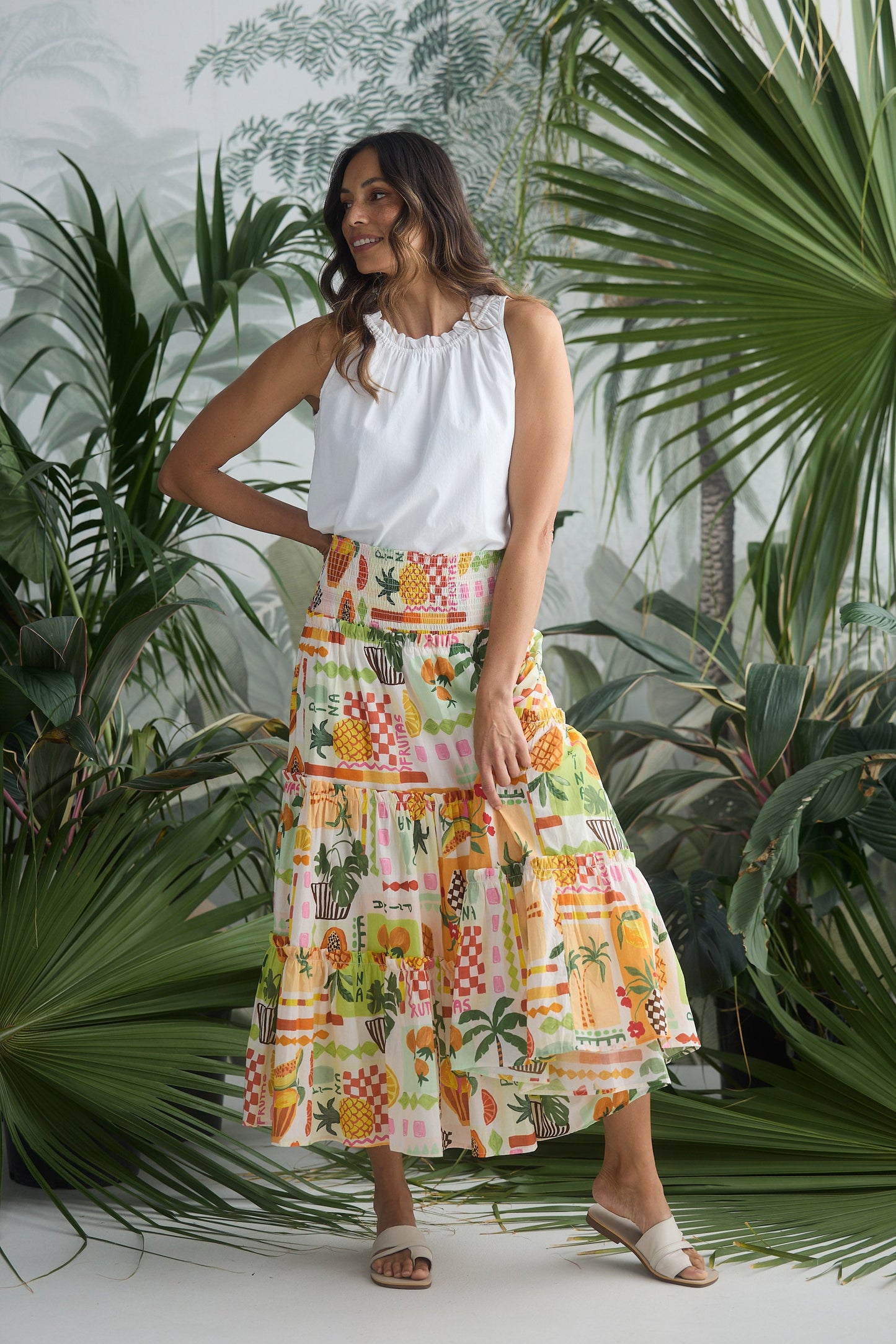 Woman wearing a white top and colorful patterned skirt standing among tropical plants.