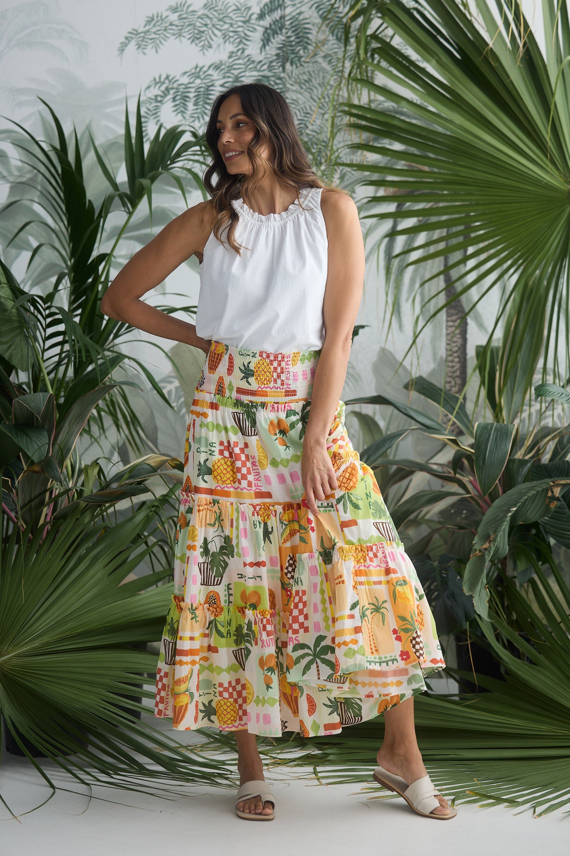 Woman wearing a white top and colorful patterned skirt standing among tropical plants.