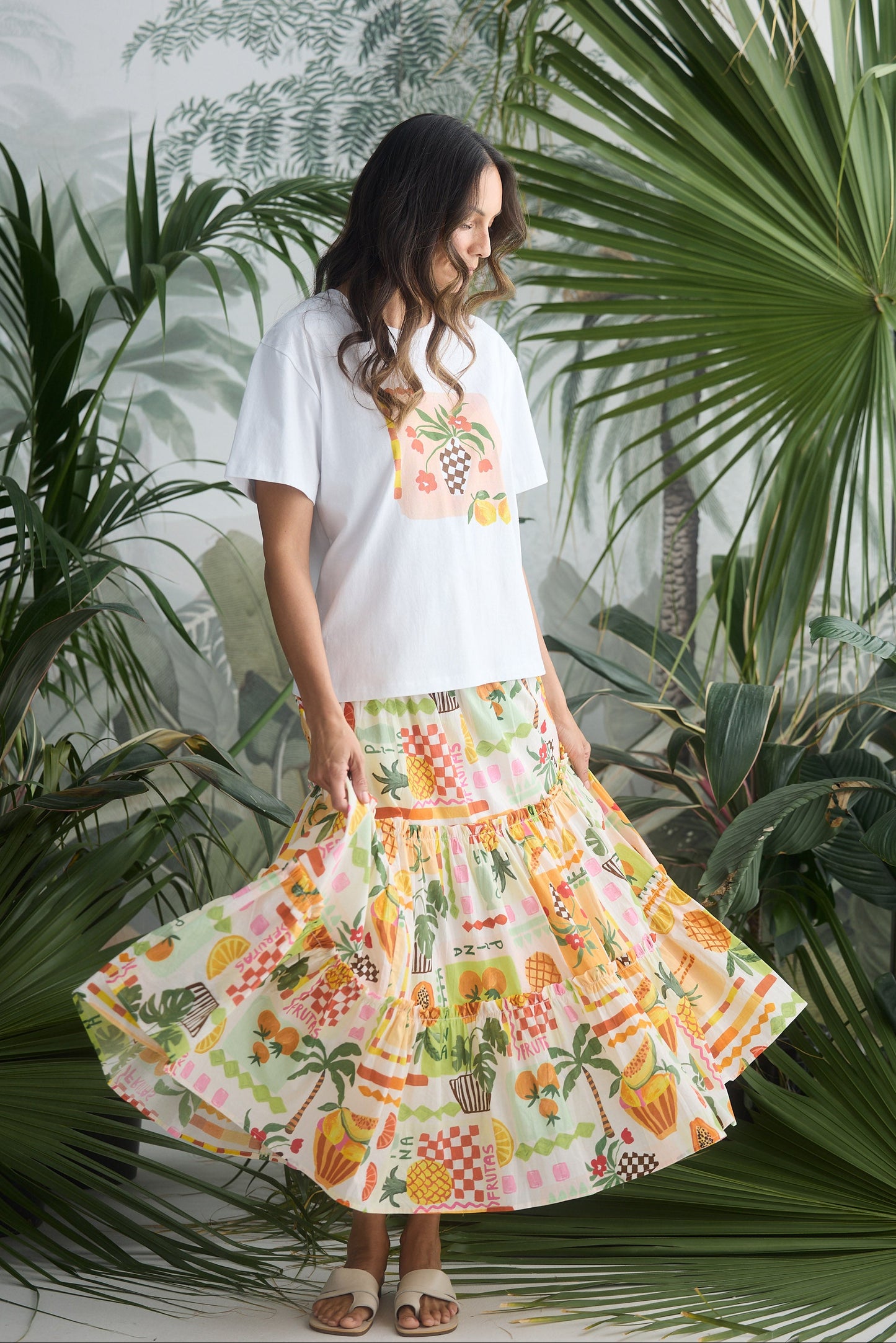Woman wearing a white t-shirt with a colorful pineapple design and a floral skirt, standing among tropical plants.