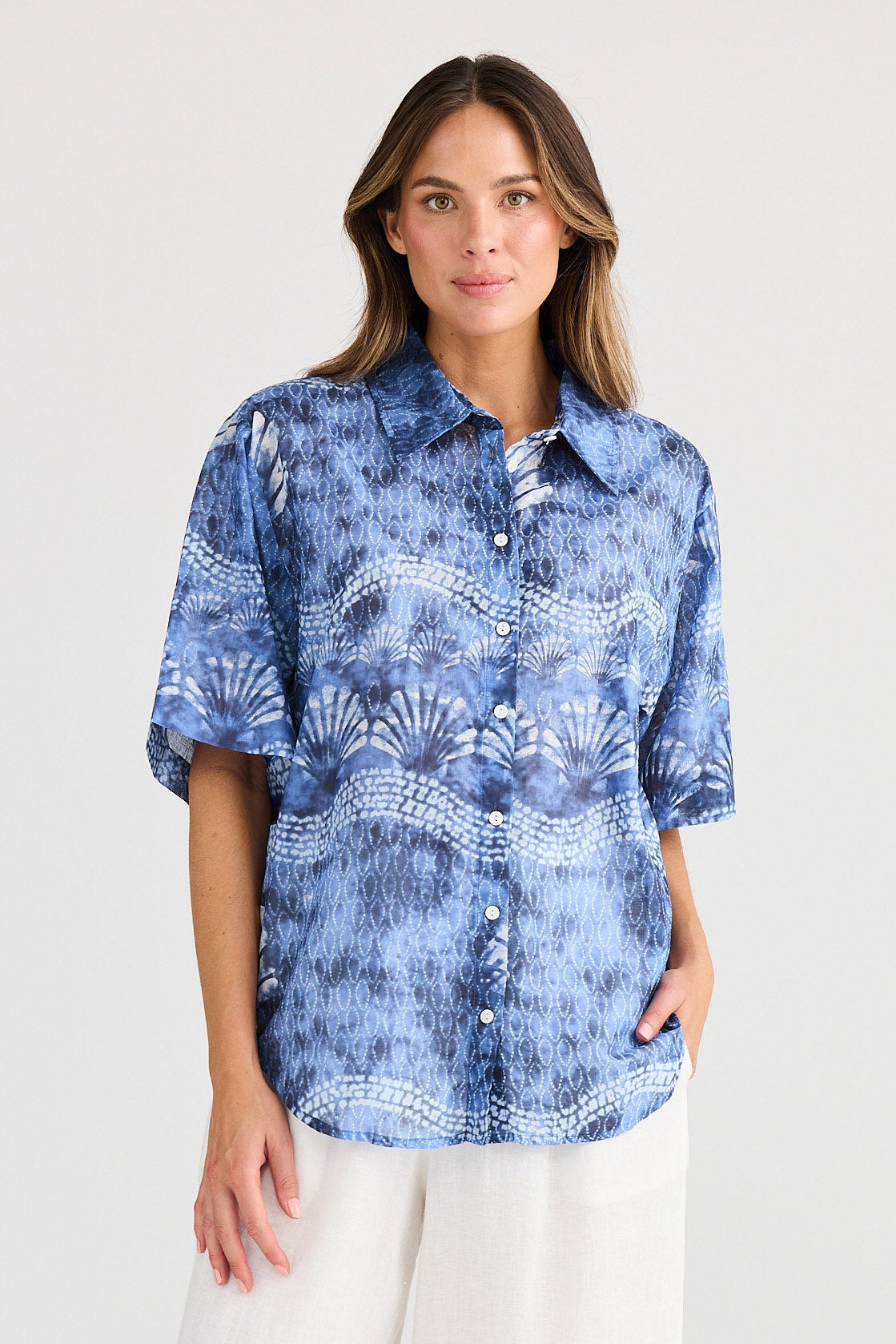 Woman wearing a blue patterned shirt and white pants on a plain background