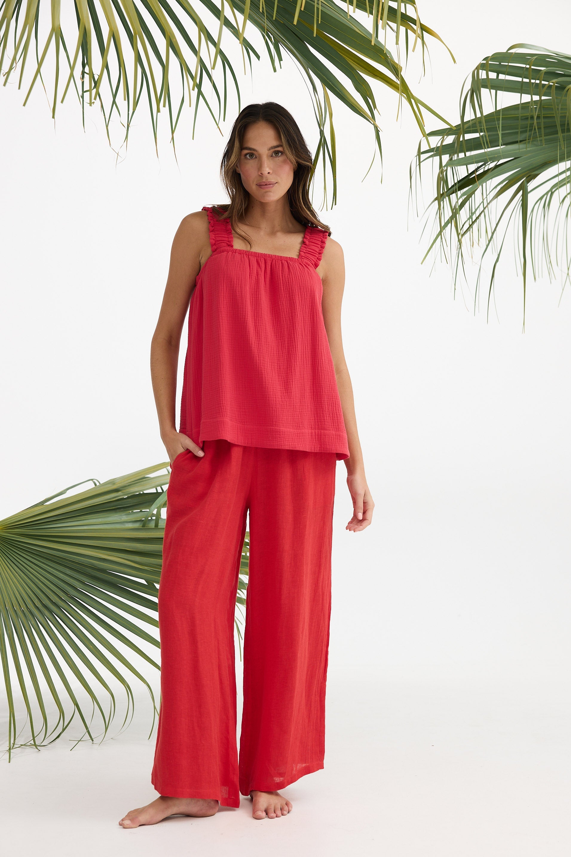 Woman wearing a red sleeveless top and wide-leg pants standing against a white background with green palm leaves.