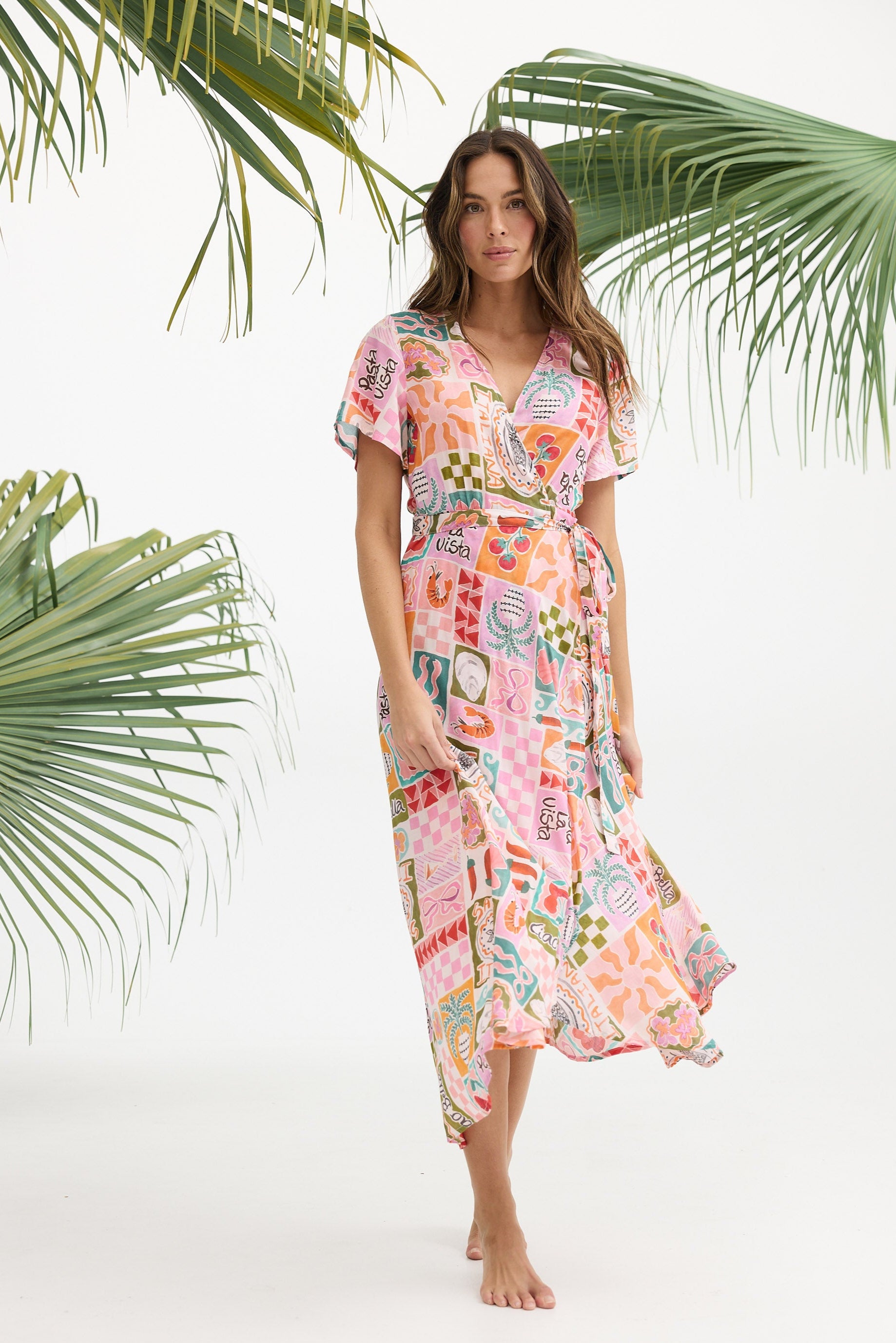 Woman wearing a colorful patterned dress standing against a white background with palm leaves.