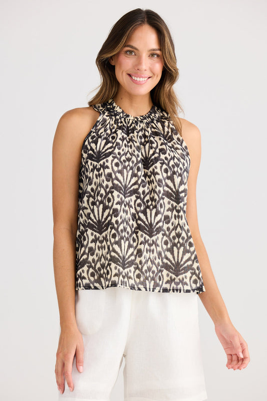 Woman wearing a patterned sleeveless top and white pants on a plain background