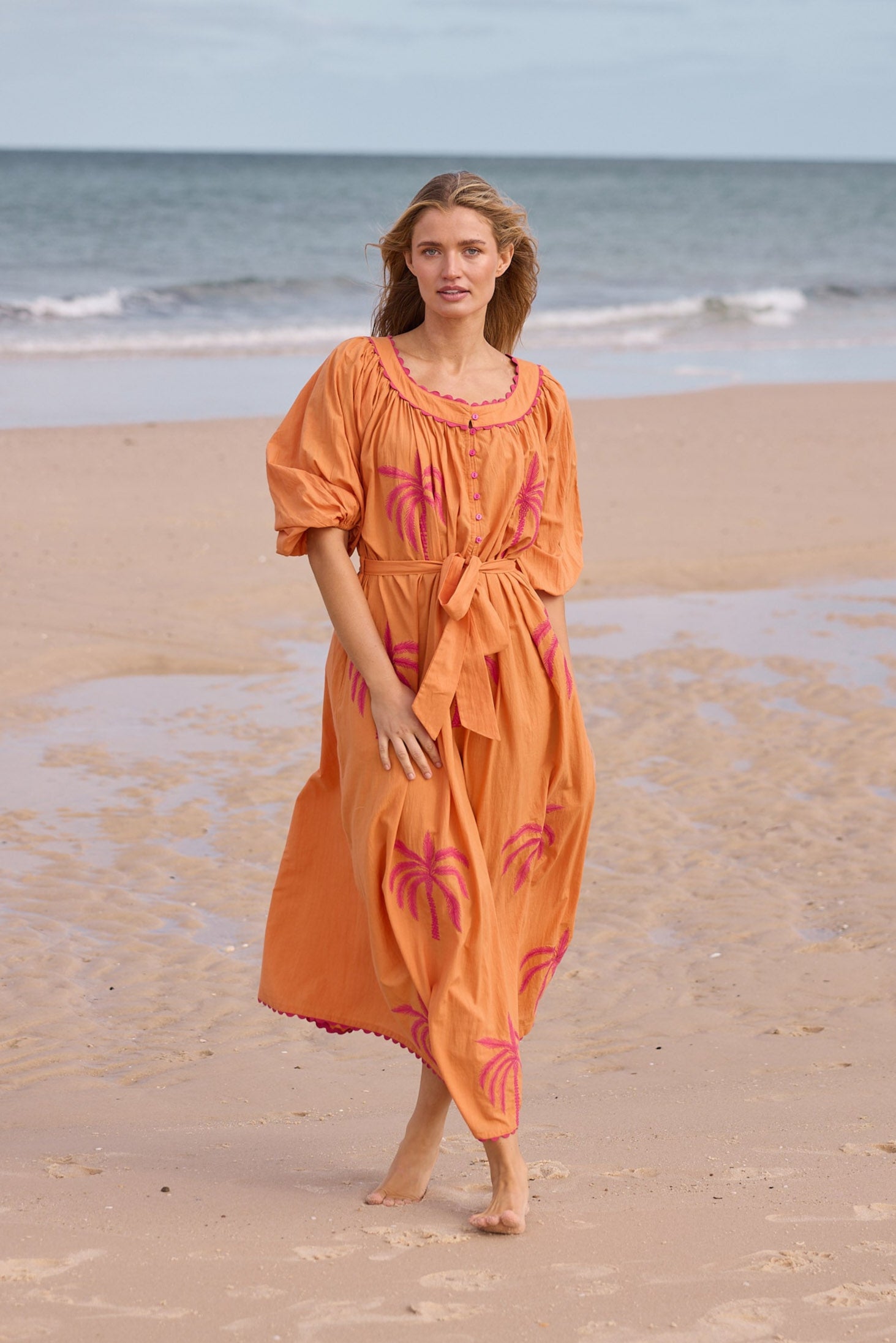 Woman in an orange dress with pink floral patterns walking on a beach.
