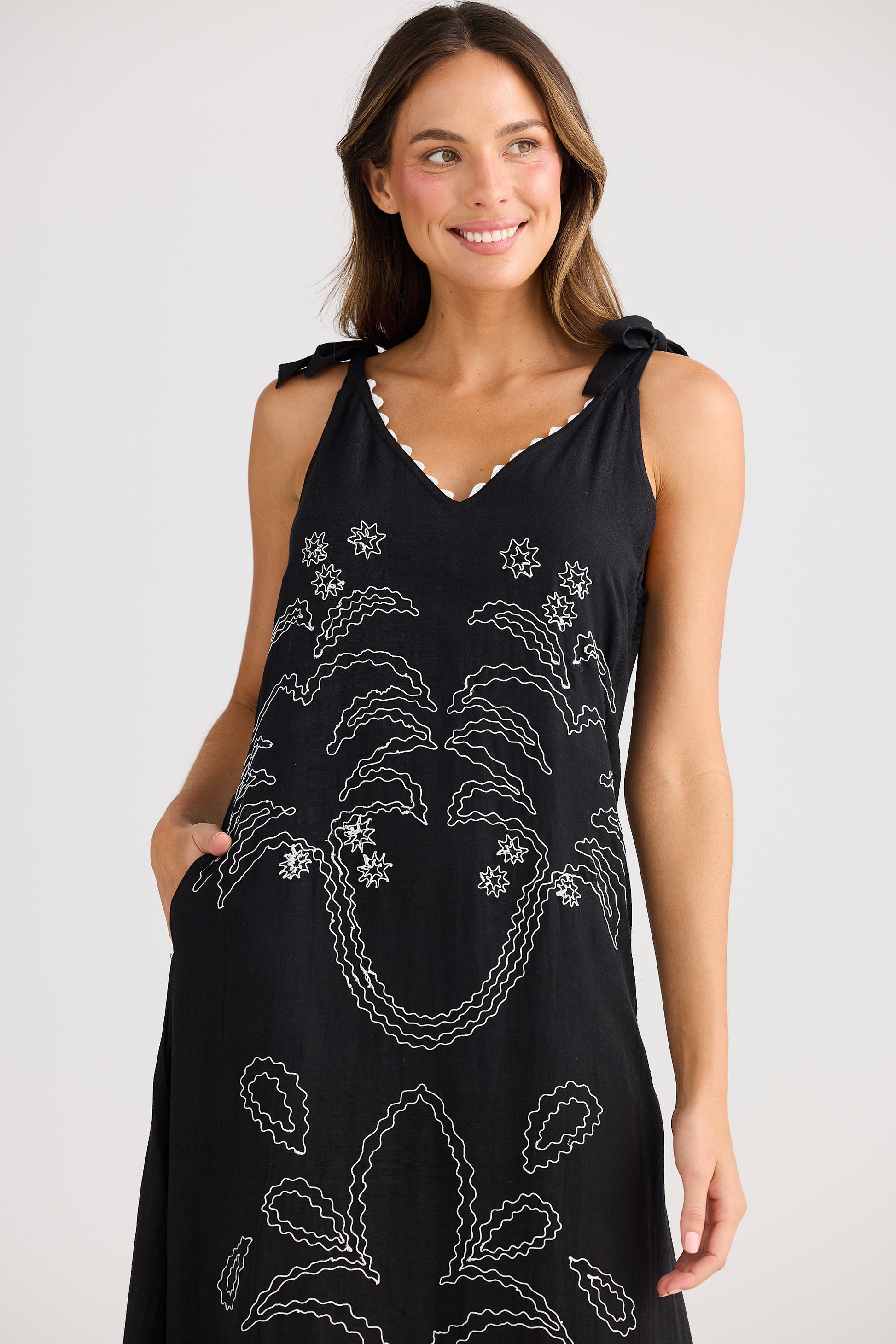 Woman wearing a black sleeveless dress with white floral embroidery on a plain background