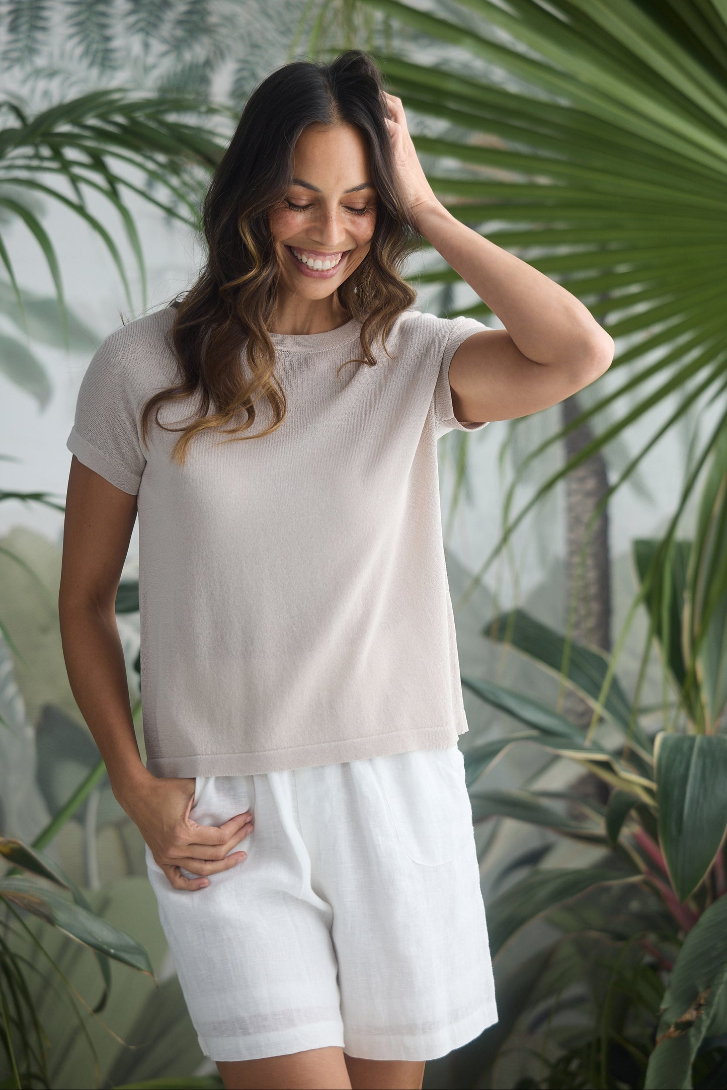 Woman wearing a beige top and white shorts standing in front of green foliage