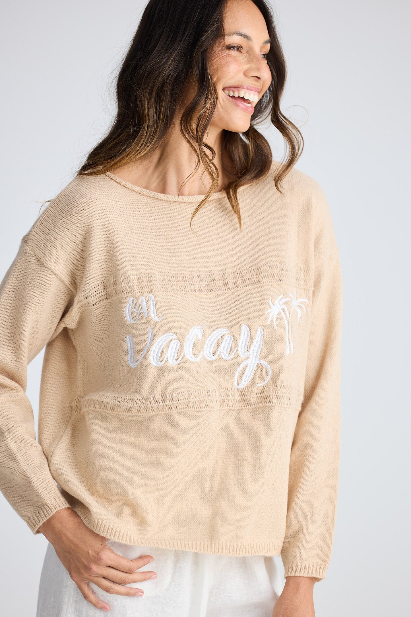 Woman wearing a beige sweater with 'on vacay' text on a plain background