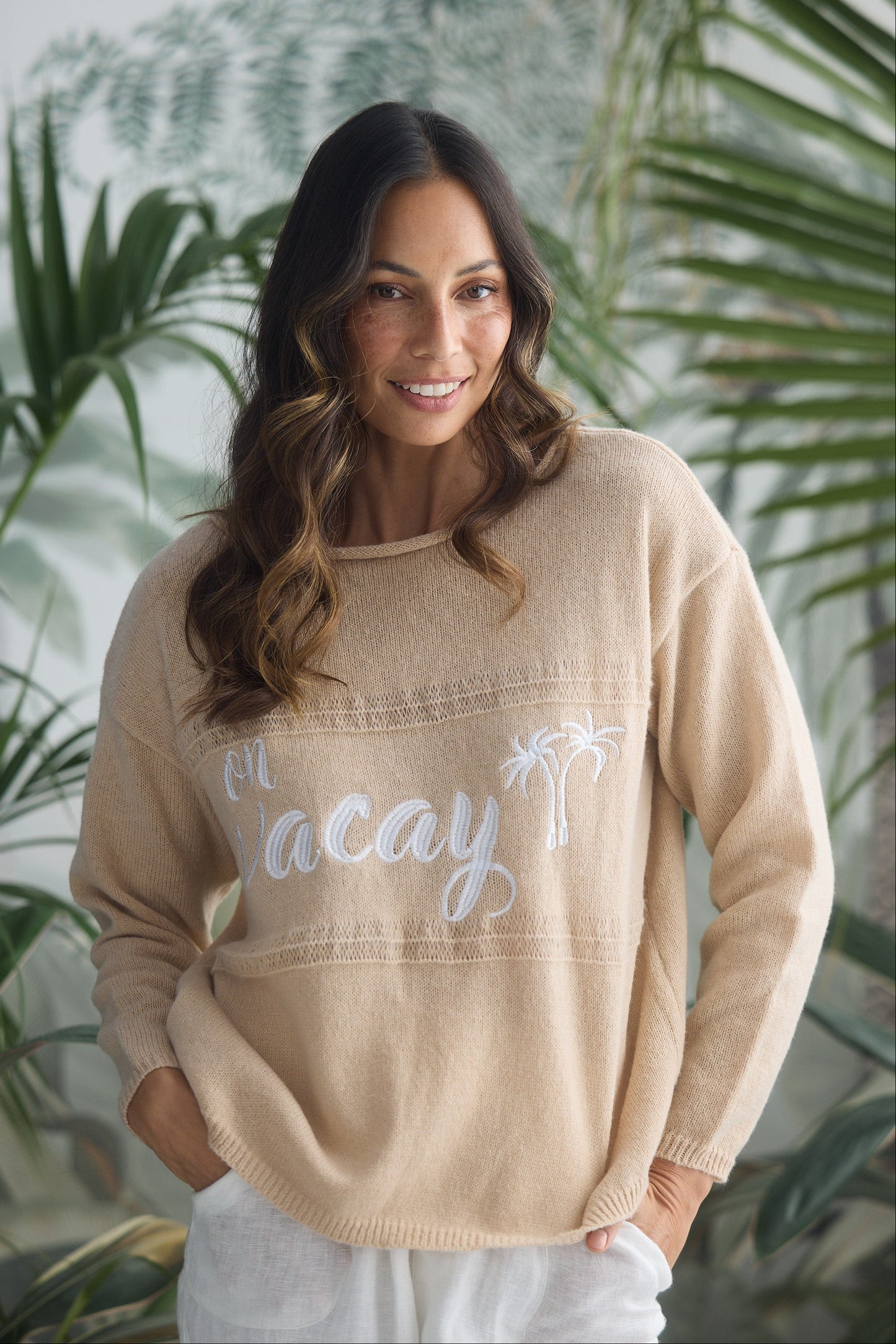 Woman wearing a beige sweater with 'On Vacay' text and palm tree design, standing in front of green foliage.