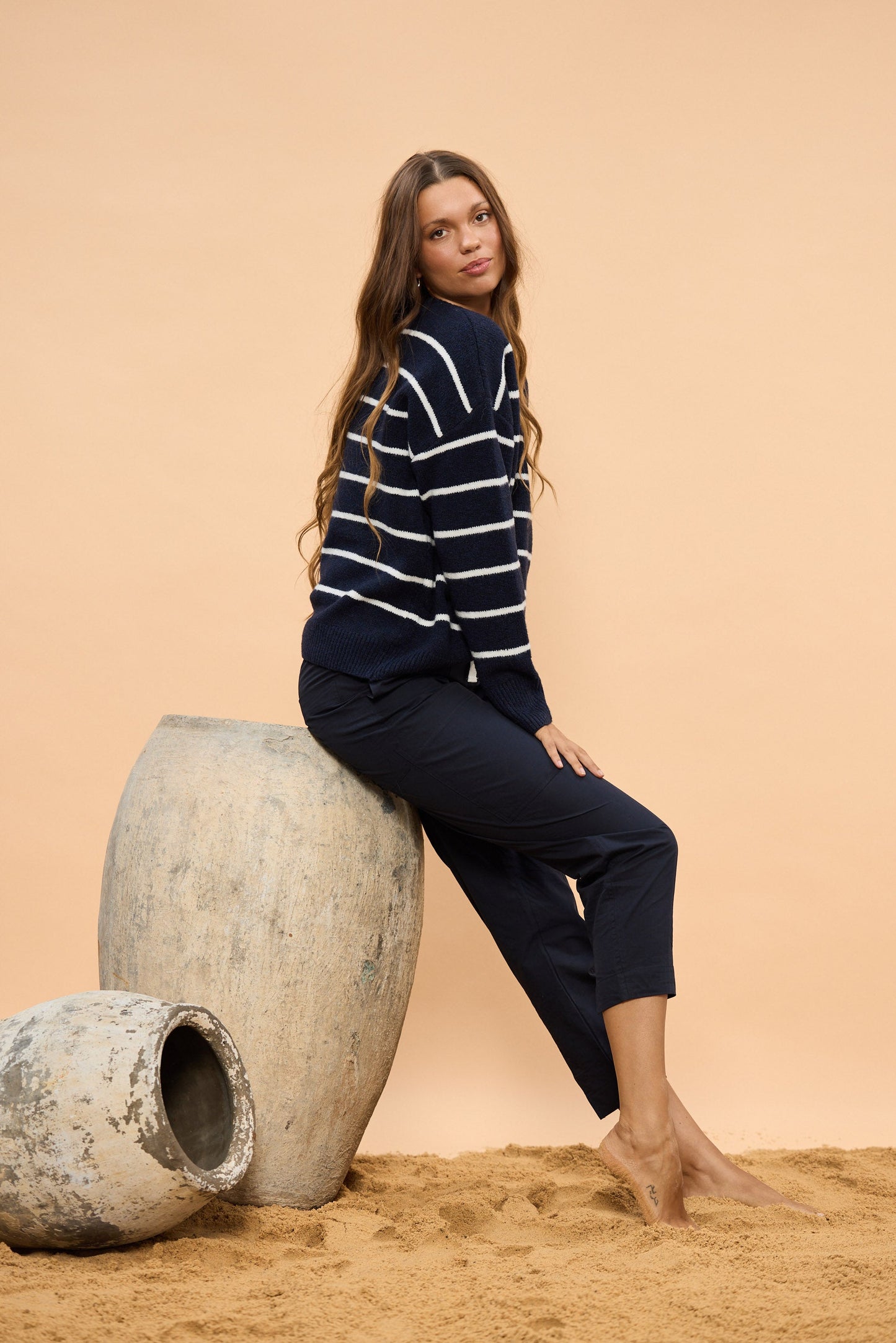 Woman in a striped sweater and pants sitting on a large pot object against a beige background