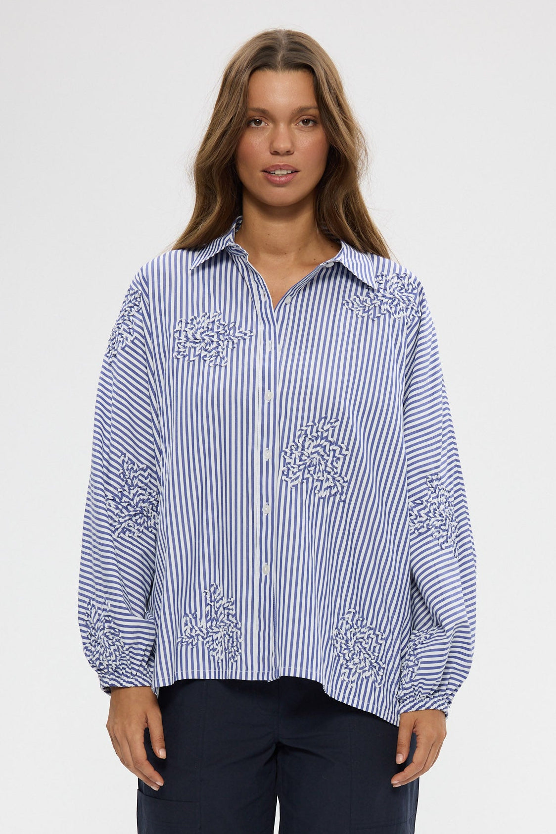 Woman wearing a blue and white striped shirt with embroidery, paired with navy pants on a white background.