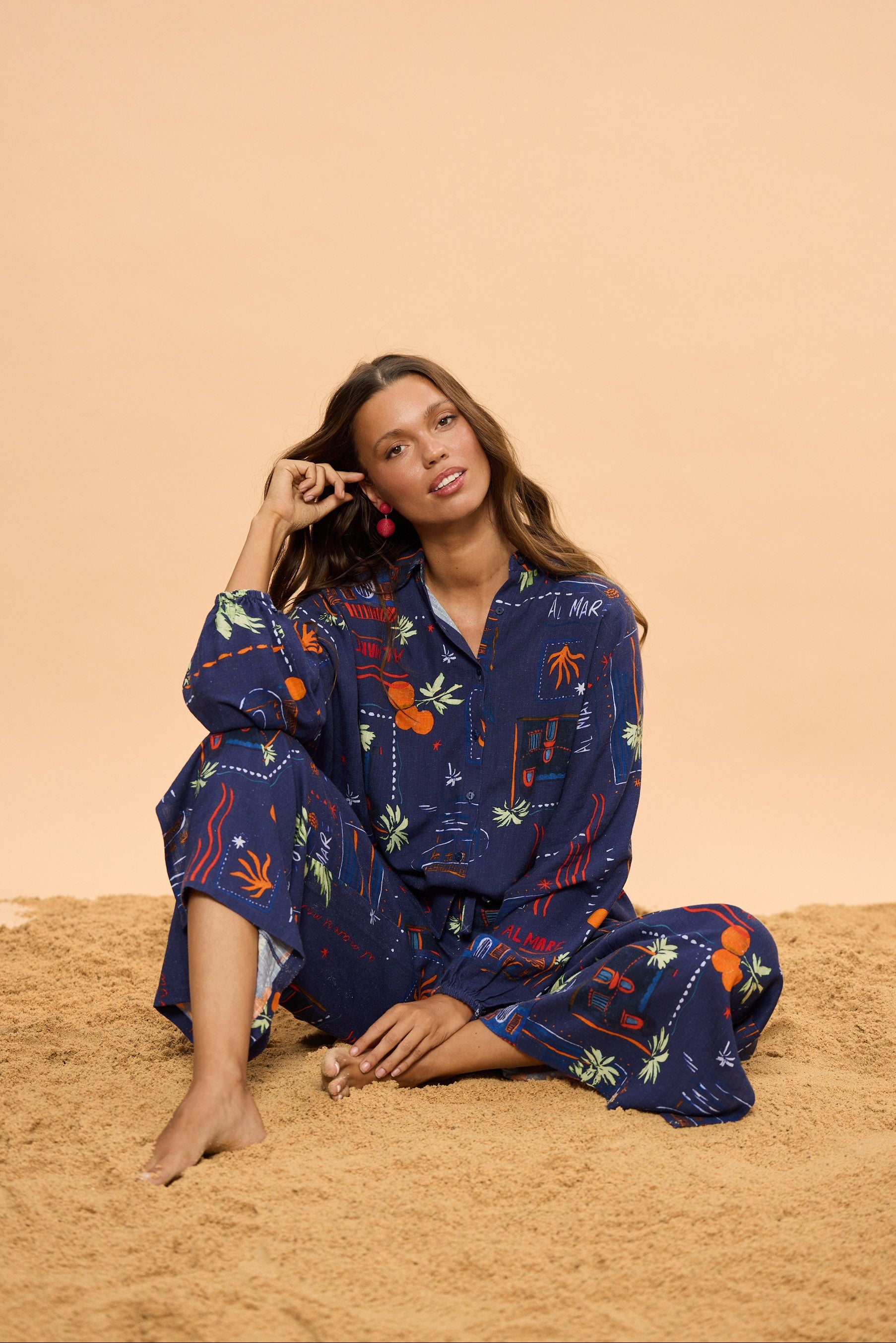 Woman wearing a blue floral shirt and pants sitting on a sand with a beige background