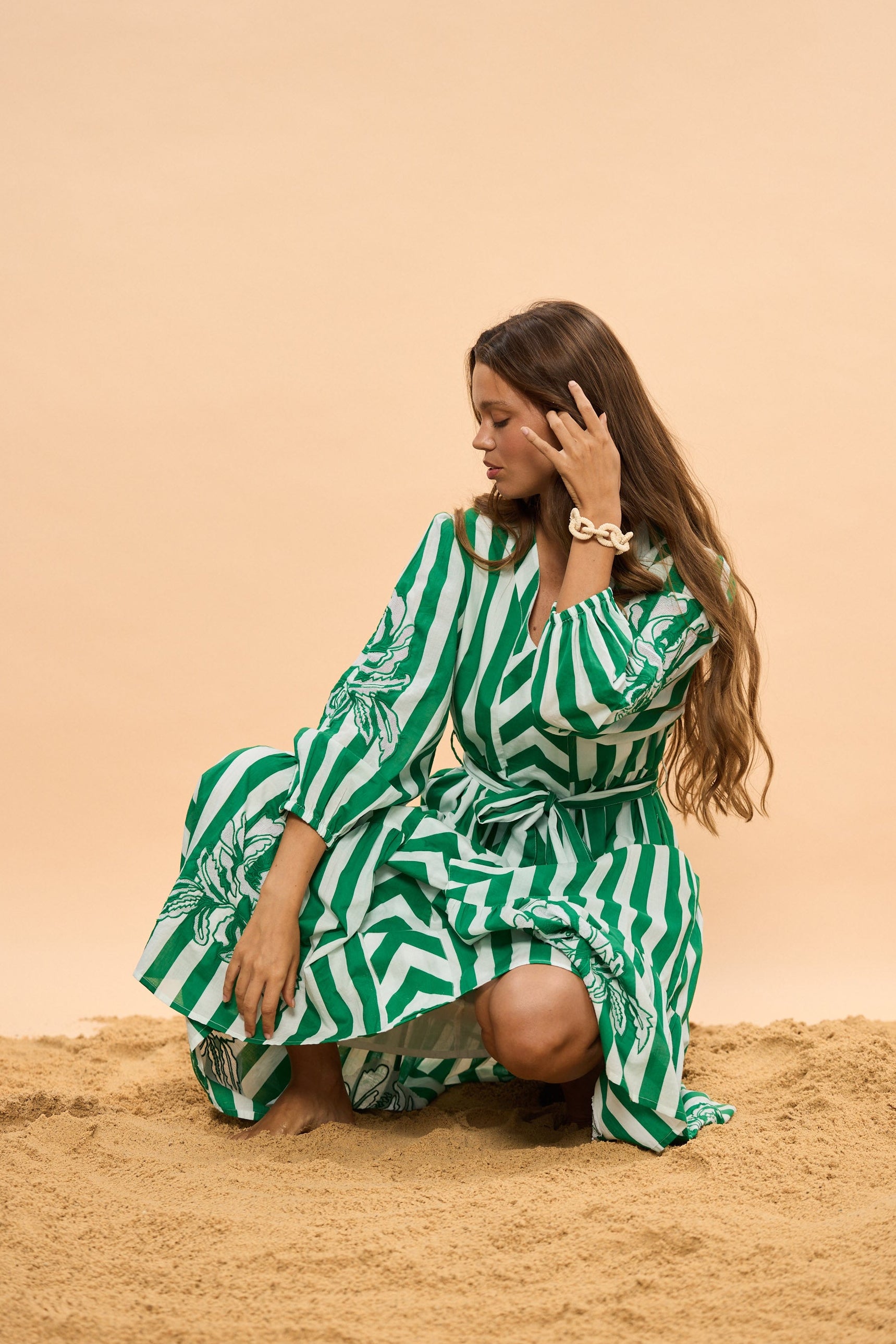 Woman in a green and white striped dress sitting on sand against a beige background