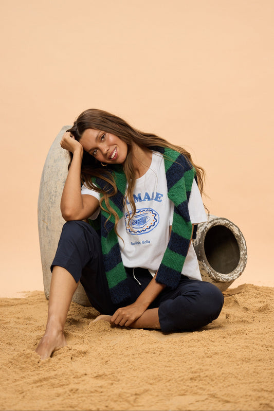 Woman sitting on sand with a beige background, wearing a white t-shirt with graphic.