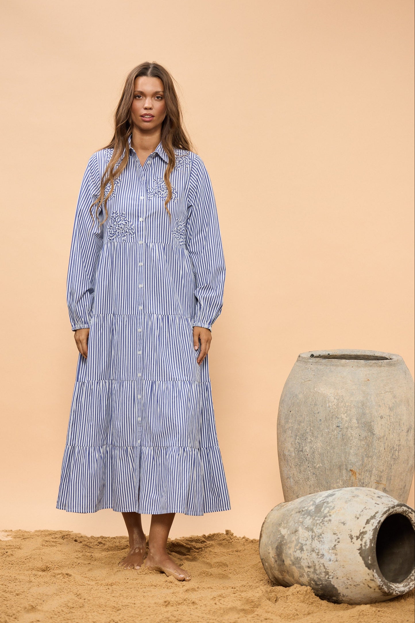 Woman in a blue striped dress standing next to large earthen pots on a beige background