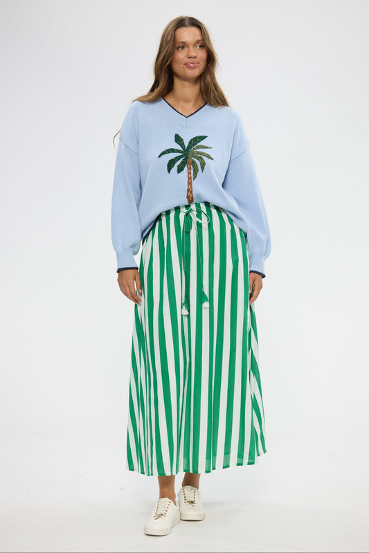 Woman wearing a light blue sweater with a palm tree design and a green and white striped skirt on a white background