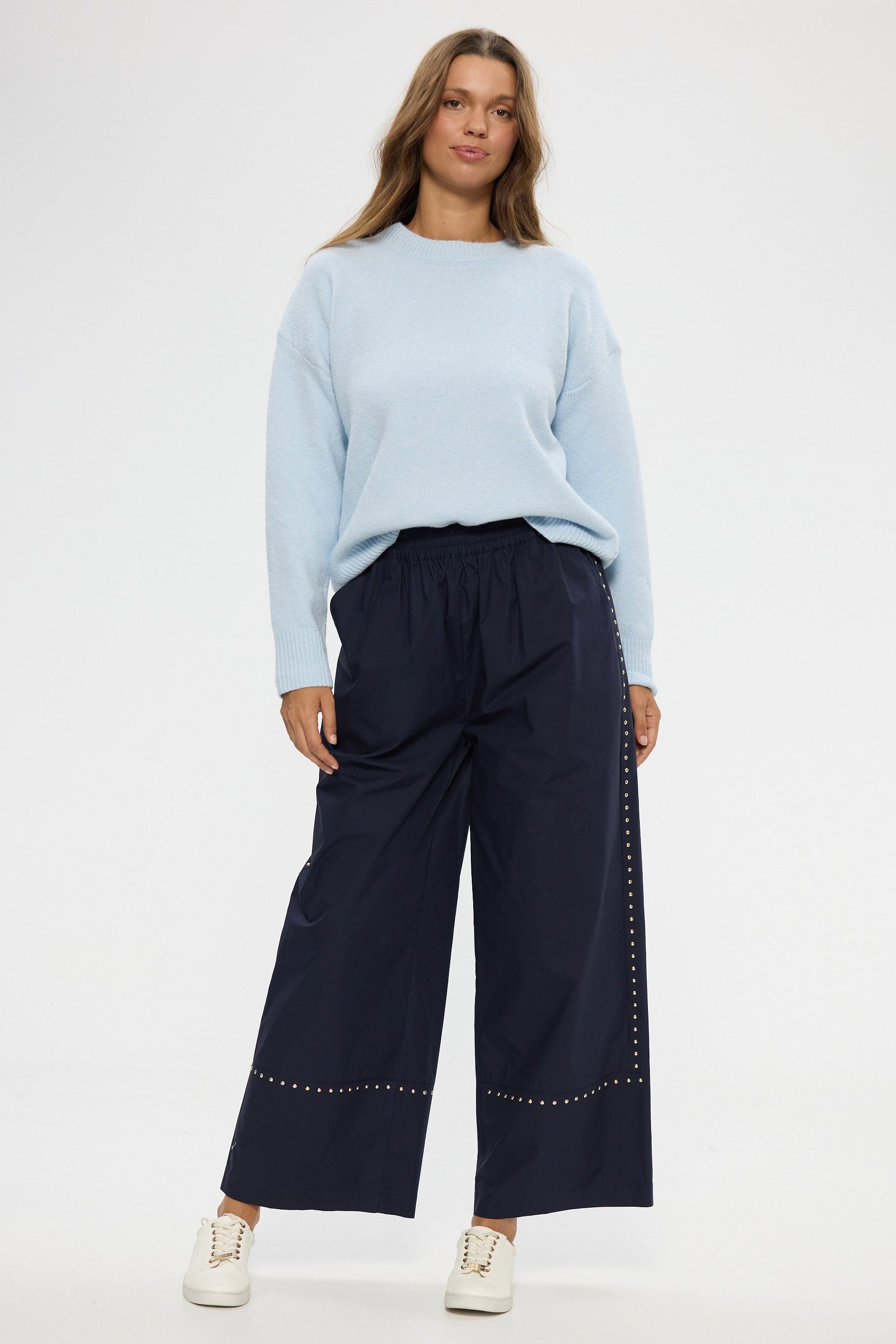 Woman wearing a light blue sweater and navy wide-leg pants with a white background