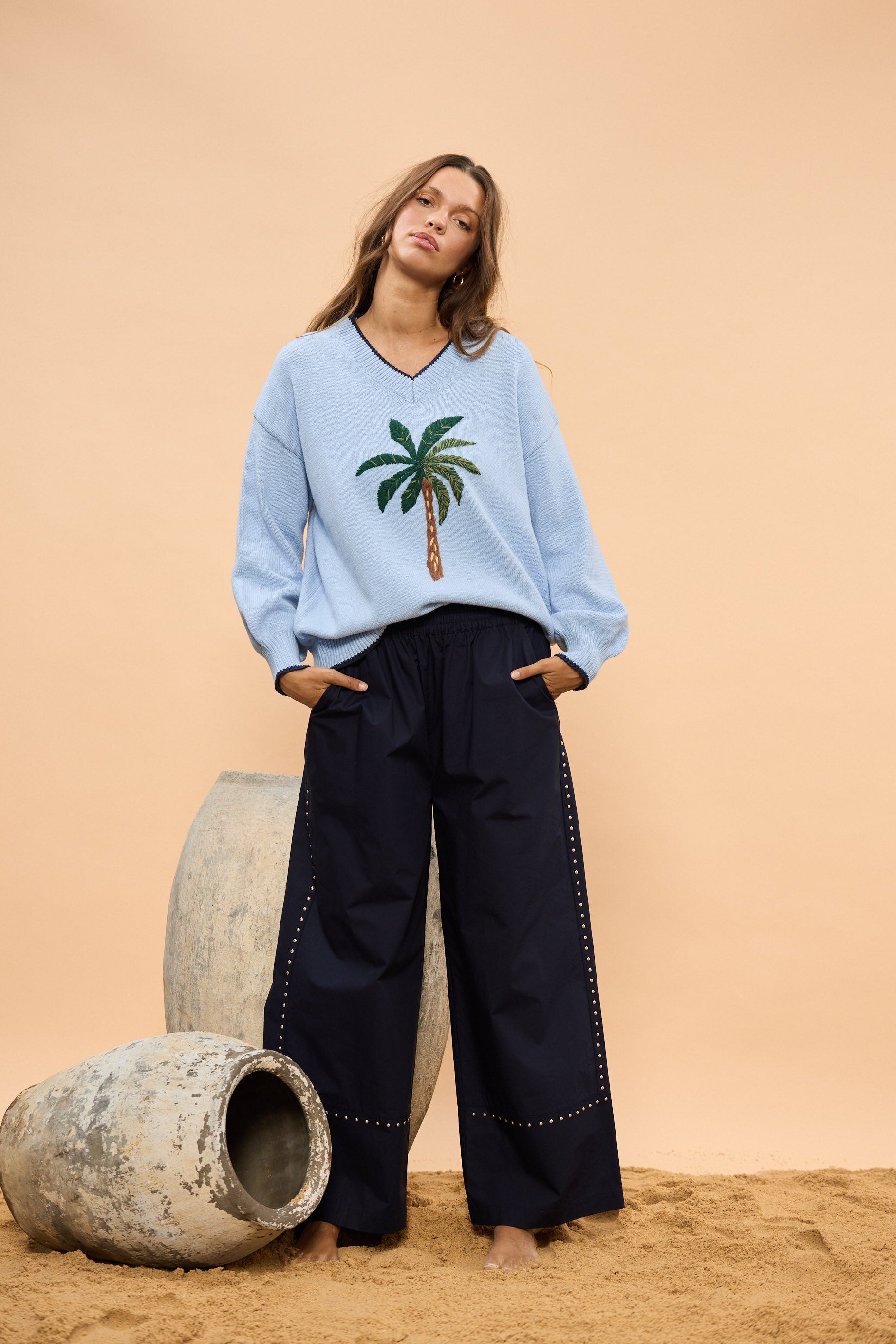 Woman wearing a light blue sweater with a palm tree design and navy wide leg pants, standing against a beige wall.