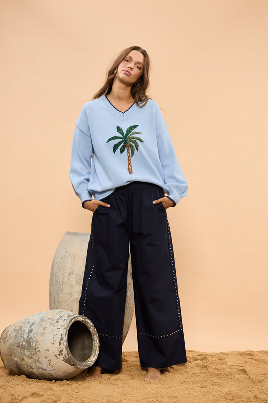 Woman wearing a light blue sweater with a palm tree design and navy wide leg pants, standing against a beige wall.