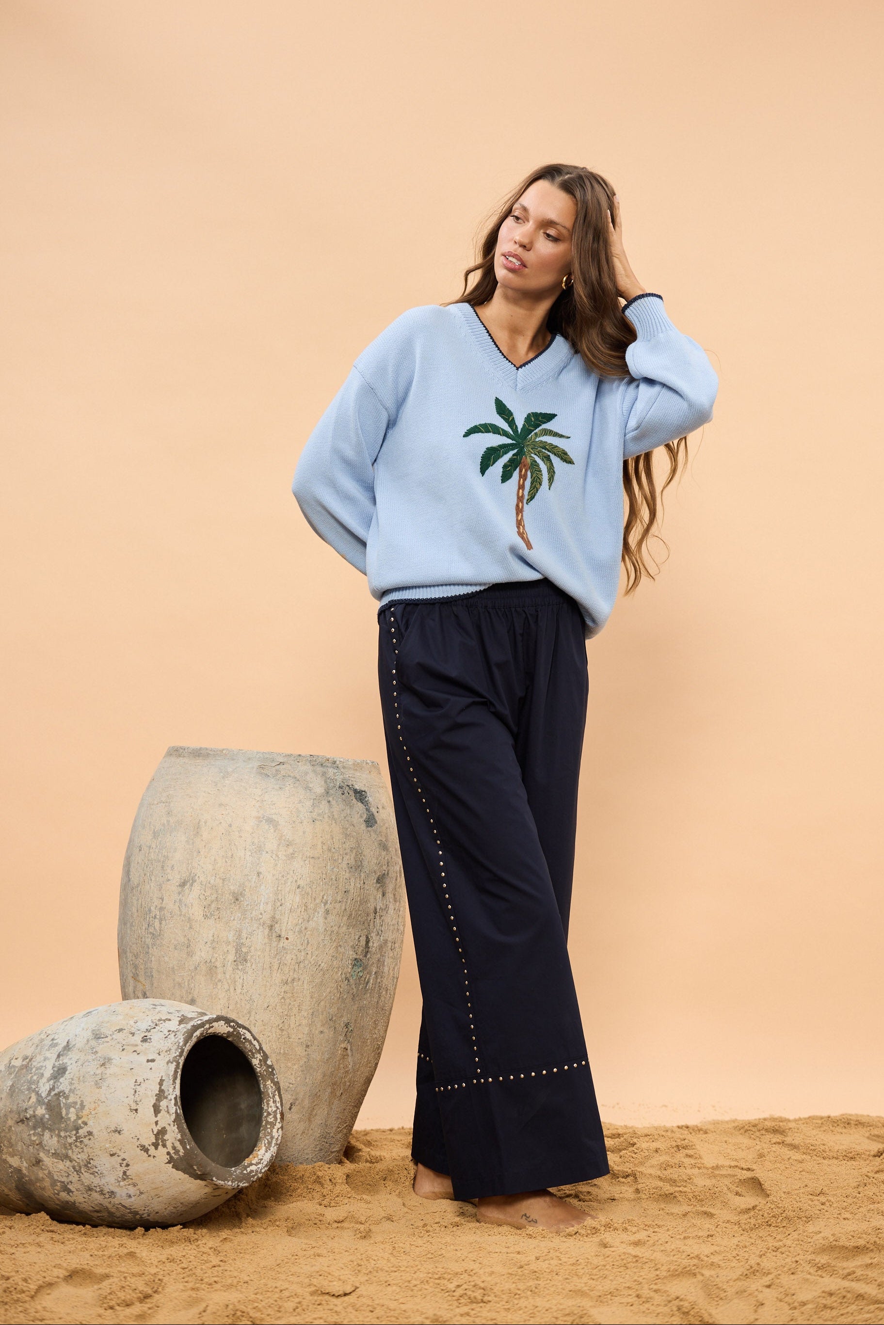 Woman wearing a light blue sweater with a palm tree design and navy pants against a beige wall.