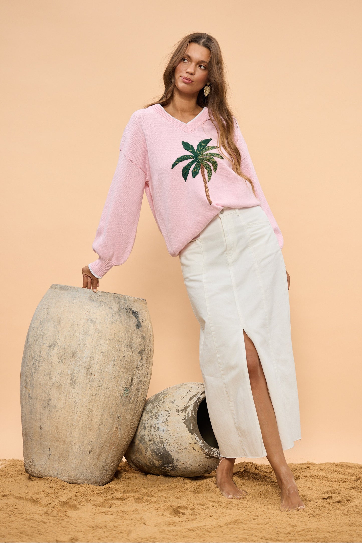 Woman wearing a pink sweater with a palm tree design, standing next to large stone vases on a beige background.