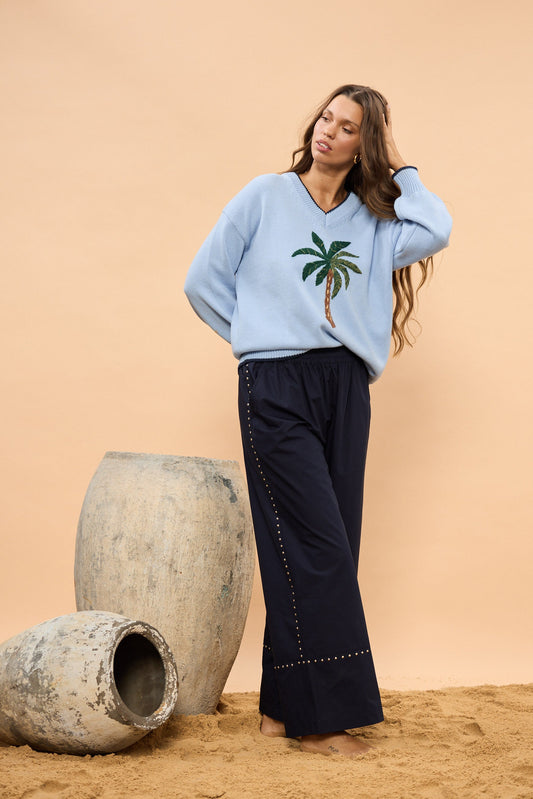 Woman wearing a light blue sweater with a palm tree design and navy pants against a beige wall.