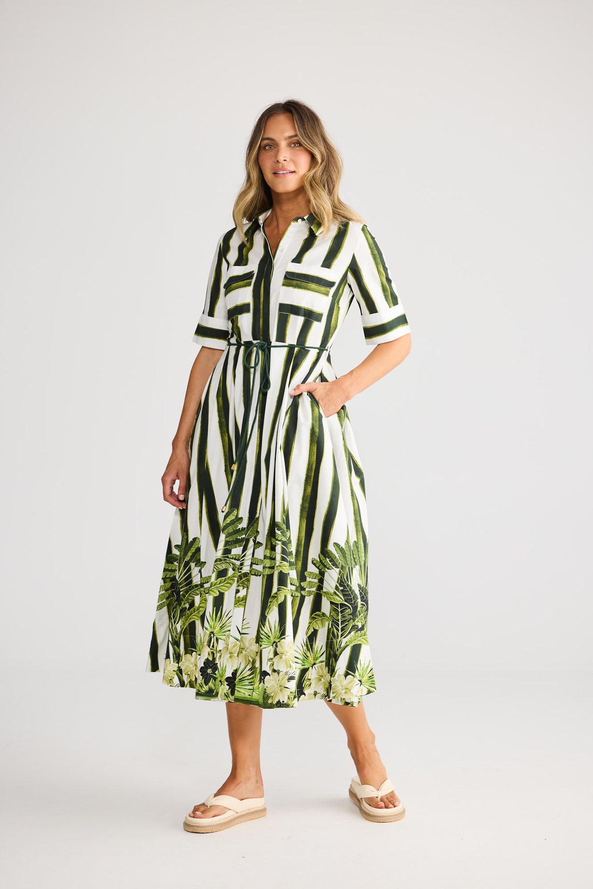 Model wearing Nalani Dress in botanica print, front view. Collared shirt dress, waist tie, green tropical leaf border print.