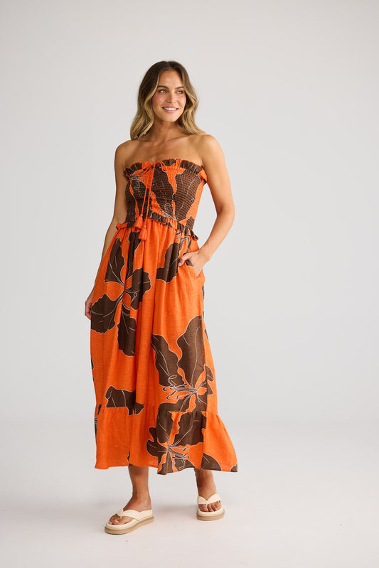 Model wearing Sorrel Dress in hibiscus breeze, front view. Strapless bodice, tiered maxi skirt, orange-brown leaf print.