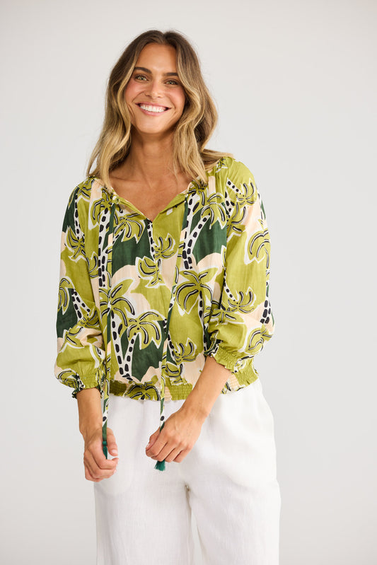 Model wearing Seaside Top in tropical deco, front view. Tasselled neck tie, raglan sleeve, green palm-tree print on cream.