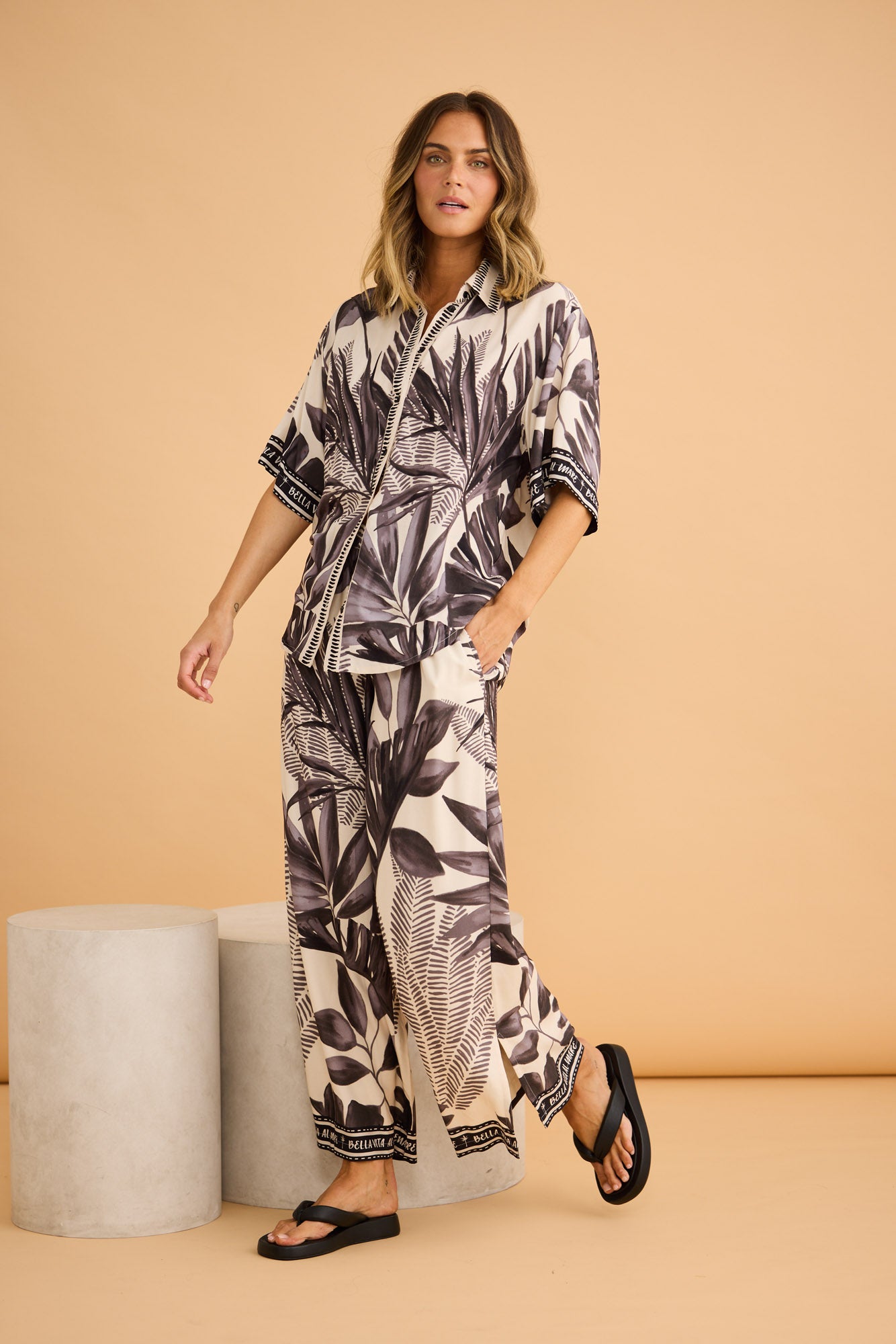 Model wearing Fly Away Pant in sunset palm, front view. Relaxed wide leg with black-and-cream palm-leaf border print.