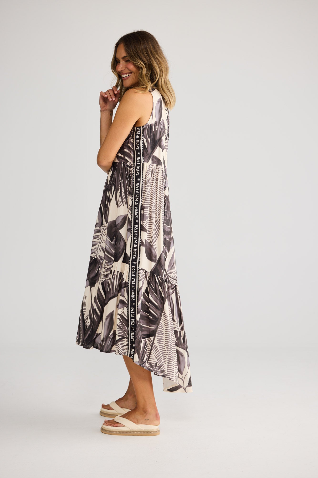 Model wearing Margot Dress in sunset palm, front view. Gathered halter neckline, tiered hi-lo skirt, tan ikat fan print.