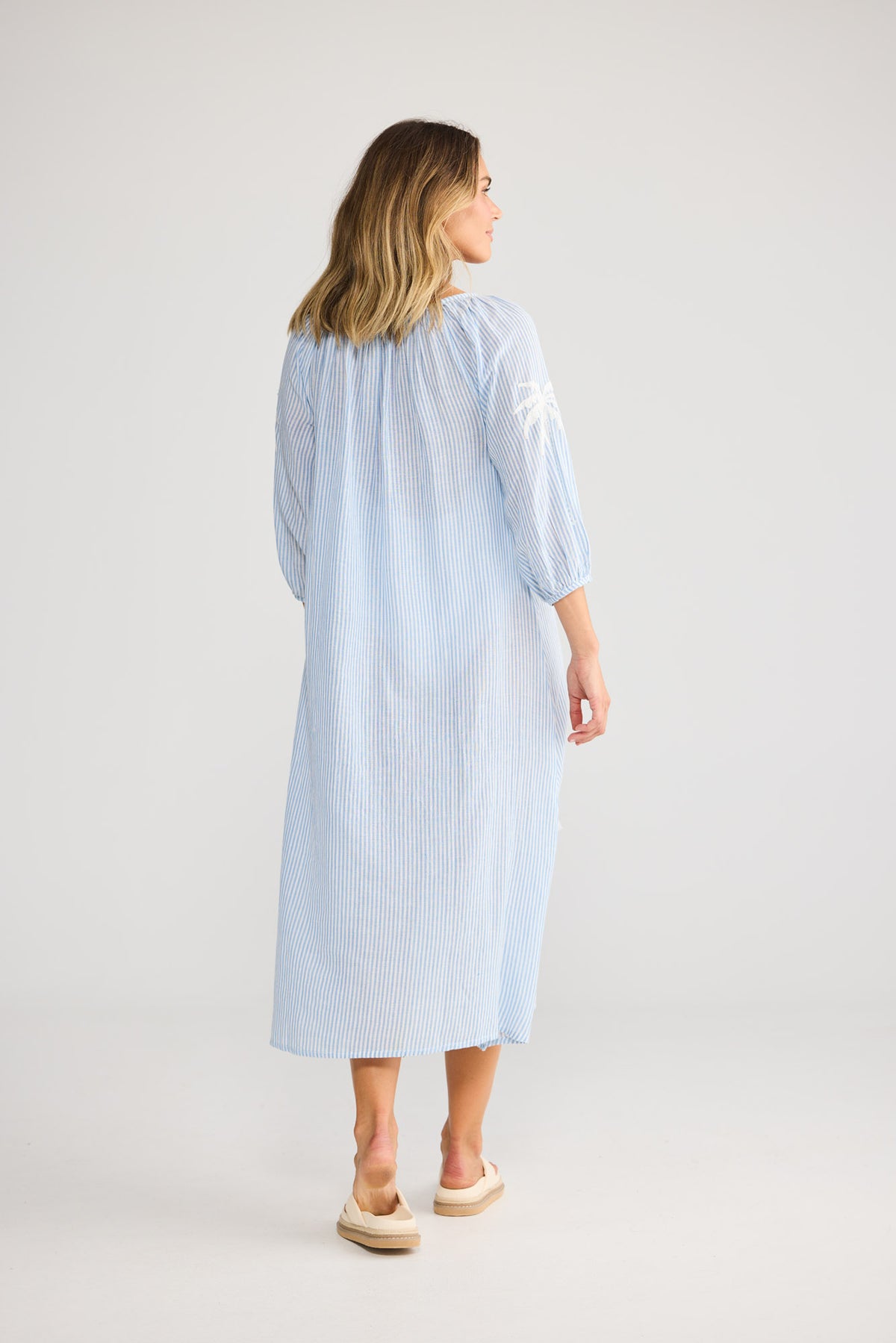 Model wearing Ohana Dress in blue stripe, front view. Neck ties, elasticated cuff sleeve, stripe with palm embroidery.