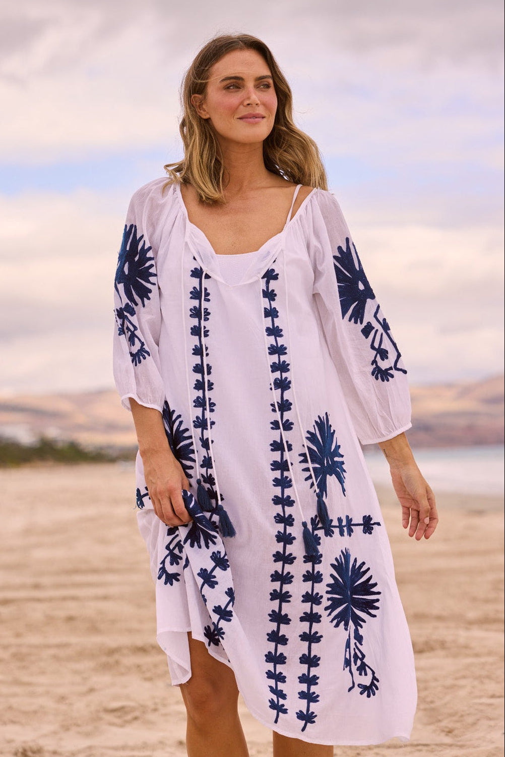 A woman wearing a white cotton cover-up dress with navy blue floral and vertical embroidered patterns, standing on a sandy beach with a cloudy sky in the background.