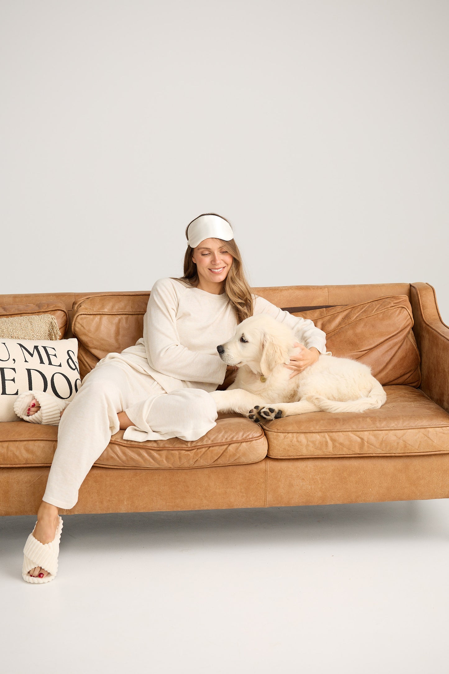 Woman in a white outfit sitting on a brown couch with two dogs, wearing a sleep mask.