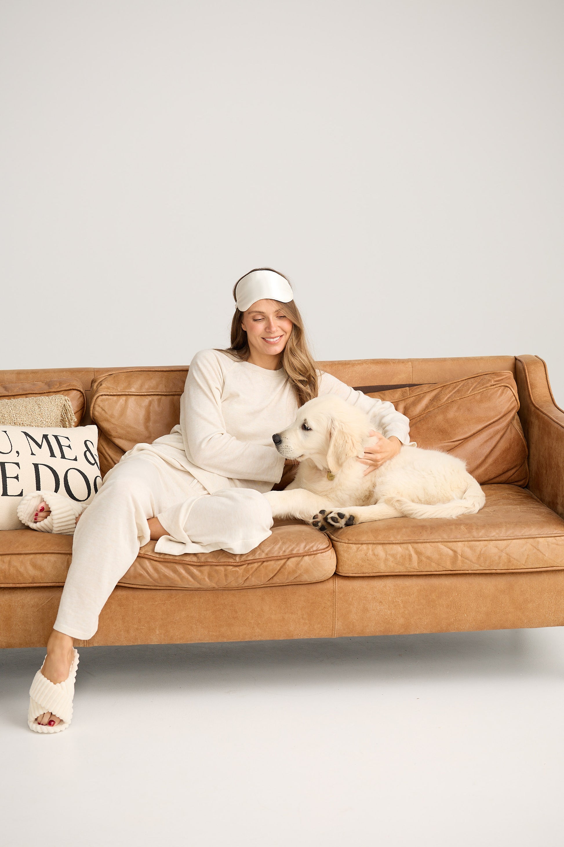 Woman in a white outfit sitting on a brown couch with two dogs, wearing a sleep mask.