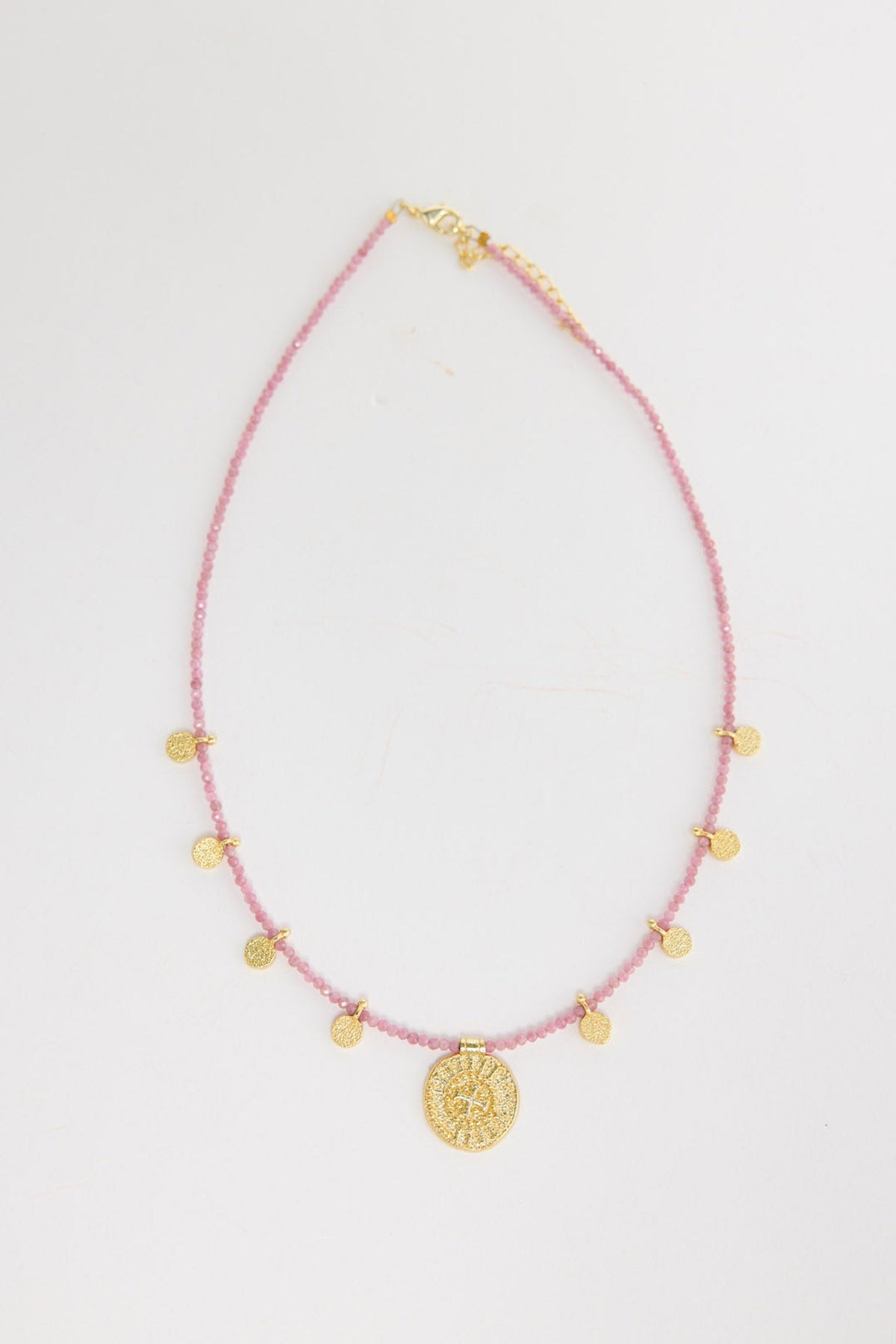Delicate pink crystal seed bead necklace with multiple small gold-tone coin charms and a larger engraved gold medallion pendant at the centre, shown flat on a white background