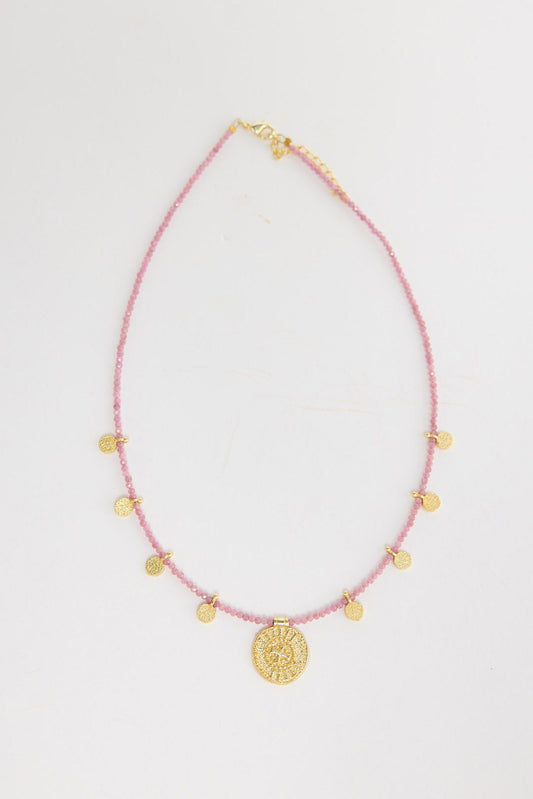 Delicate pink crystal seed bead necklace with multiple small gold-tone coin charms and a larger engraved gold medallion pendant at the centre, shown flat on a white background