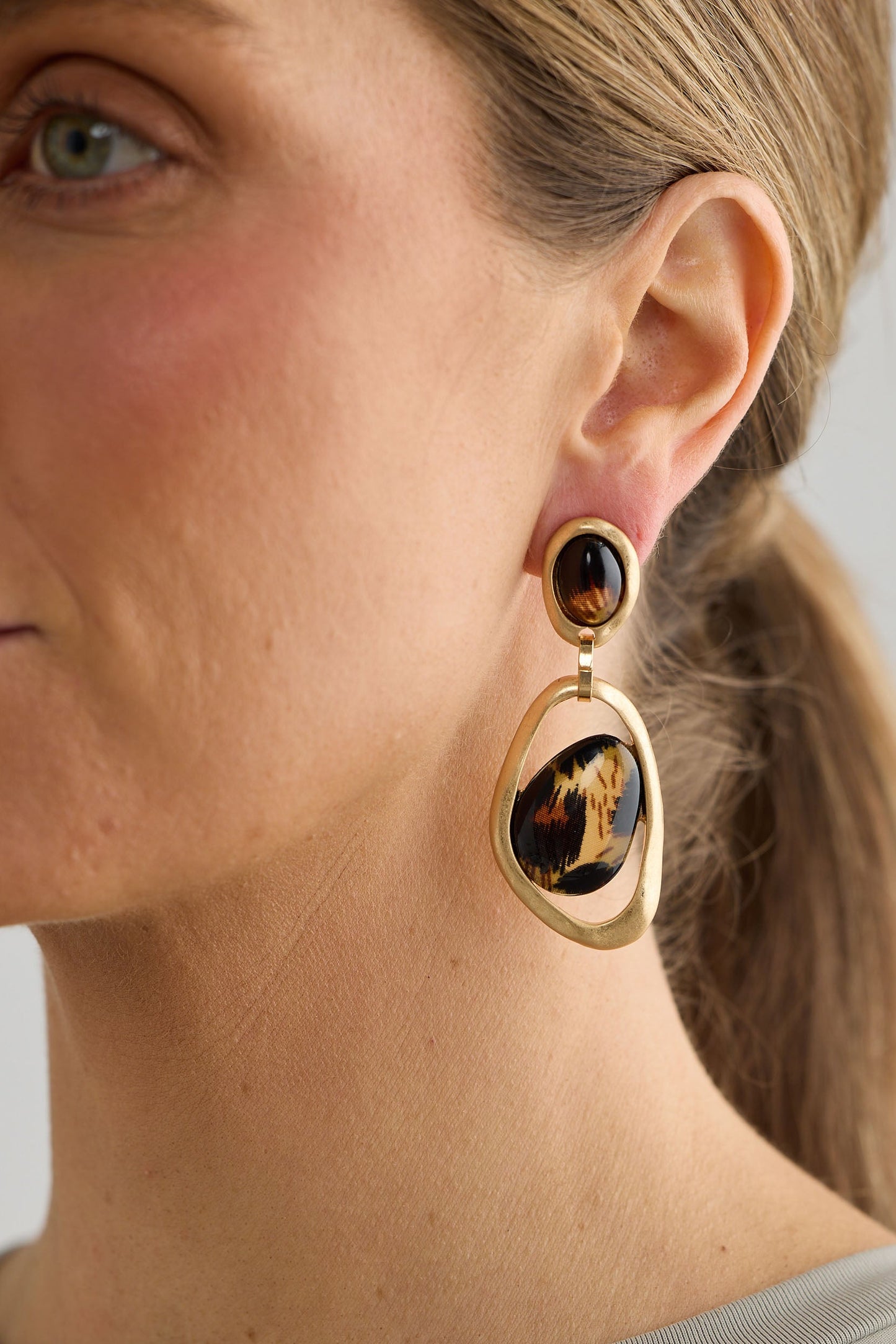 Close-up of a person wearing gold earrings with a dark gemstone on a neutral background