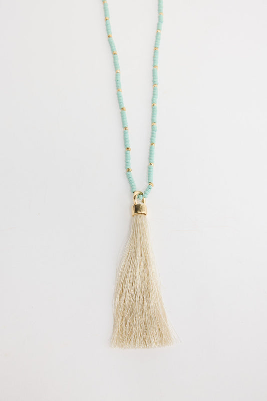 Long aqua-blue seed bead necklace with gold-tone spacer beads and a large cream silk tassel pendant with a gold cap, shown flat on a white background