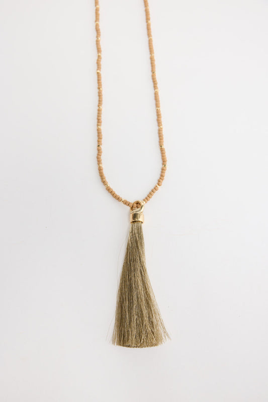 Long neutral taupe seed bead necklace with gold-tone spacer beads and a large olive-gold silk tassel pendant with a gold cap, shown flat on a white background