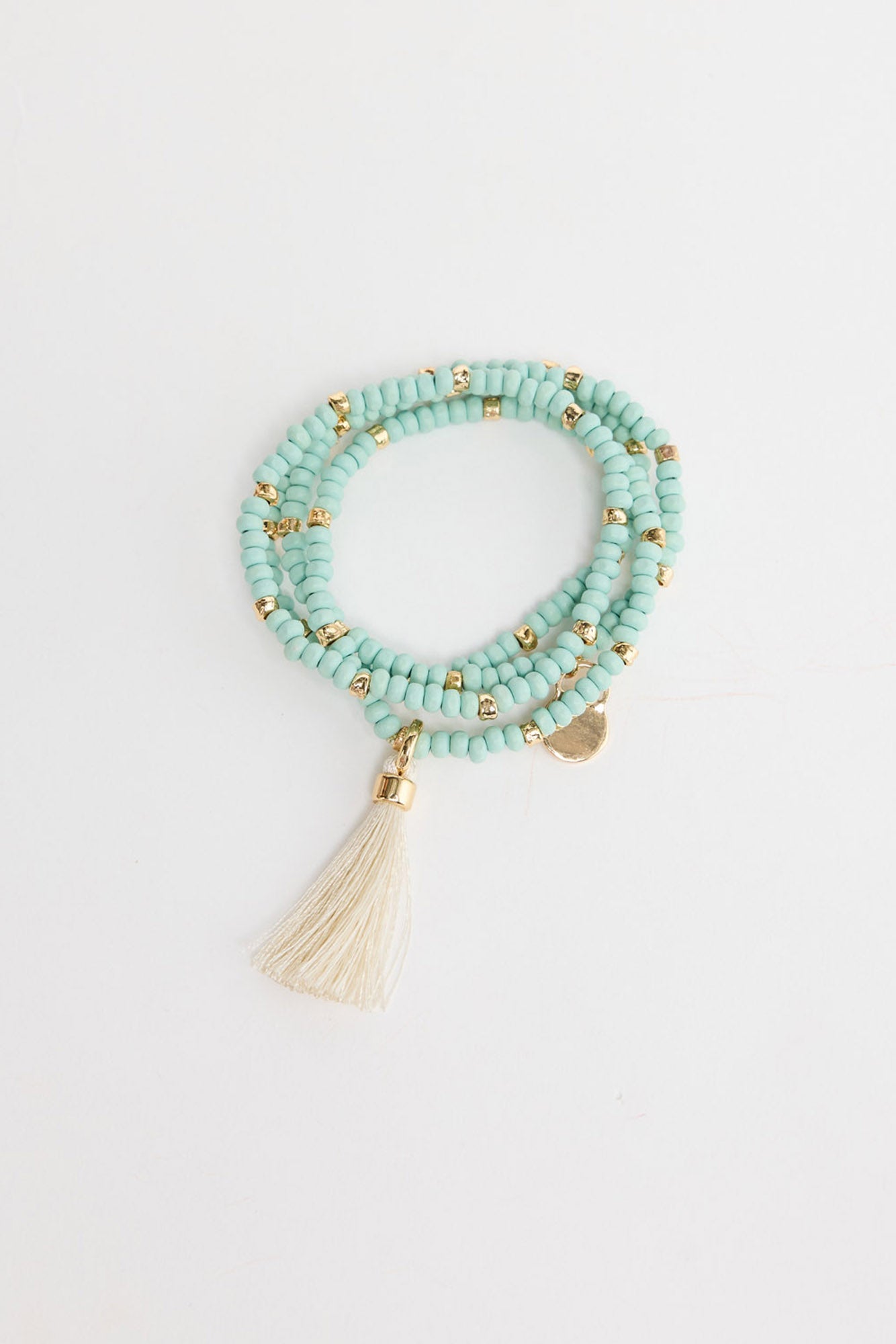 Triple-wrap stretch bracelet in aqua-blue seed beads with gold-tone accent beads, a cream silk tassel charm and a small engraved gold disc, shown coiled flat on a white background