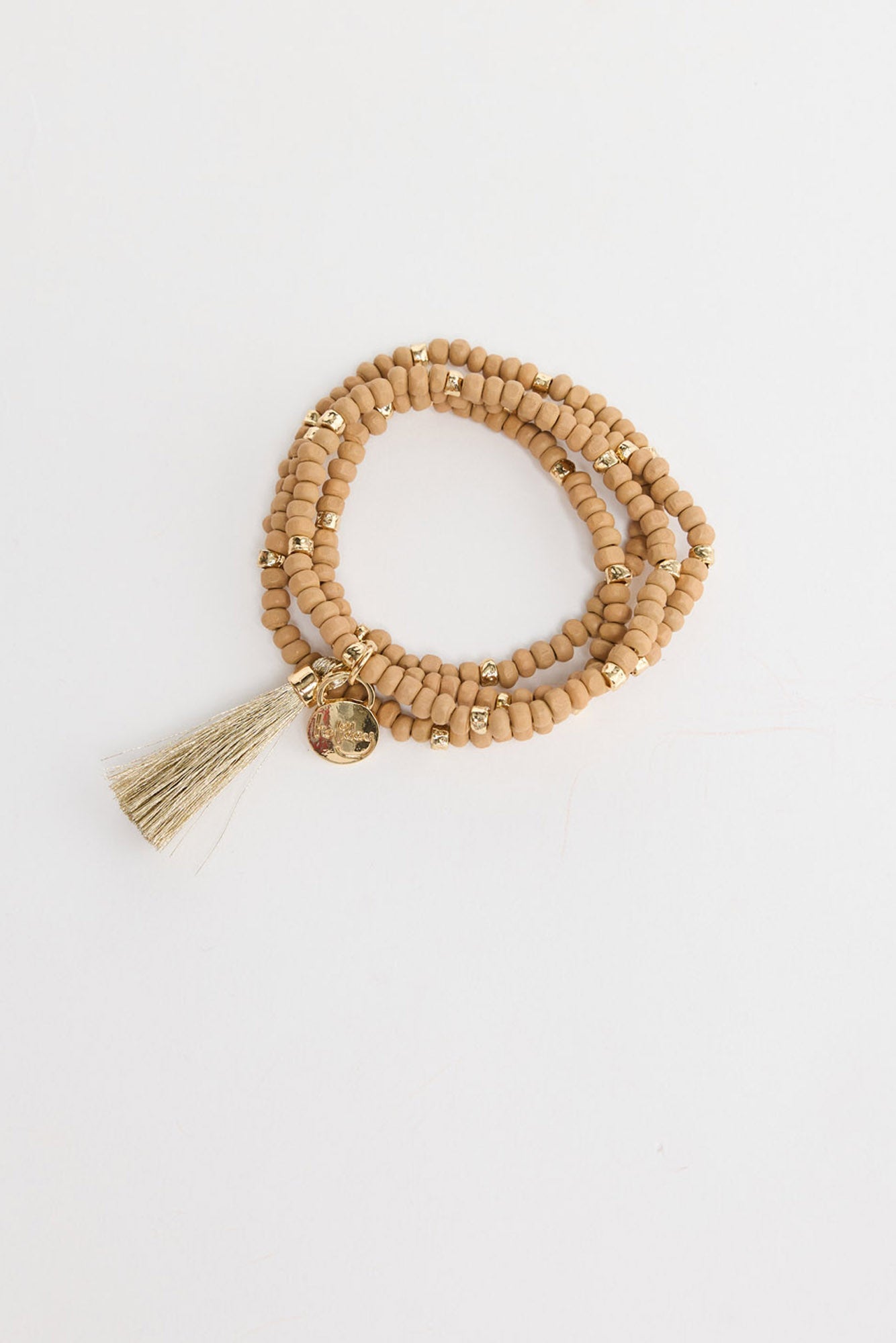 Triple-wrap stretch bracelet in neutral taupe seed beads with gold-tone accent beads, a metallic gold silk tassel charm and a small engraved gold disc, shown coiled flat on a white background