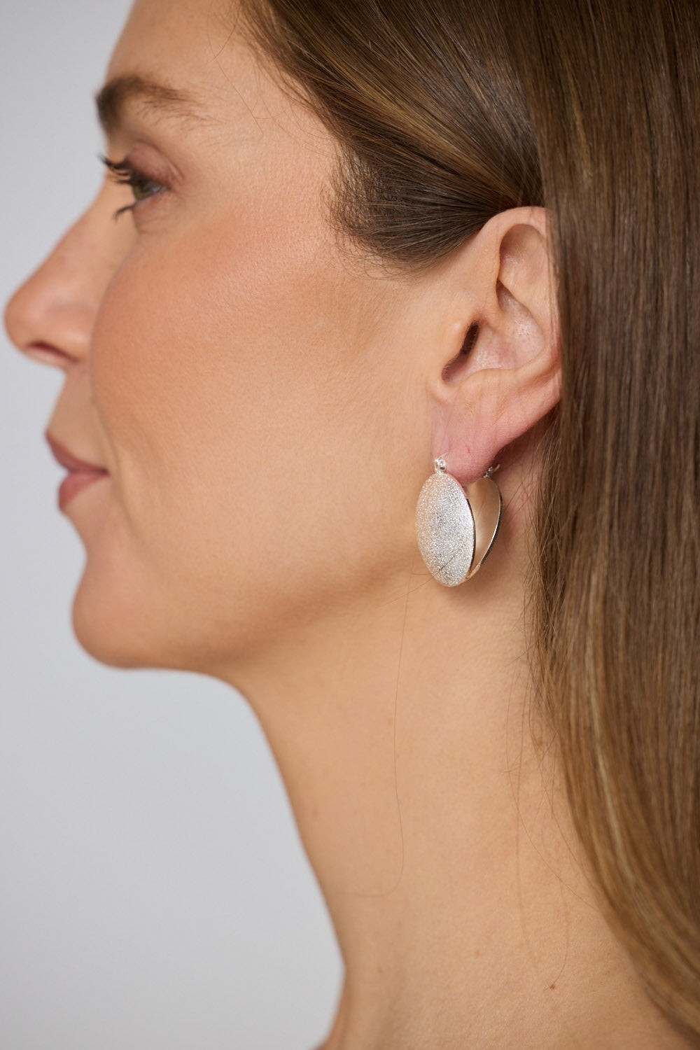 Model wearing Sue Earrings in silver with a brushed textured finish, ear detail view on neutral background