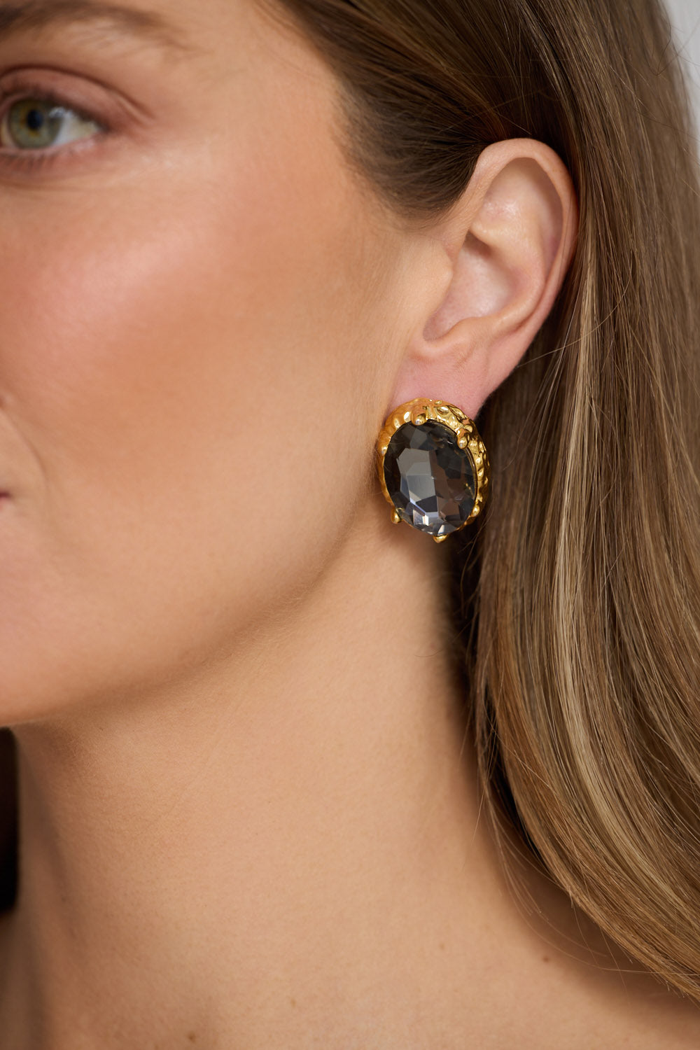 Model wearing Unice Earrings in smoke with a faceted stone set in a textured gold surround, model close-up view on neutral background